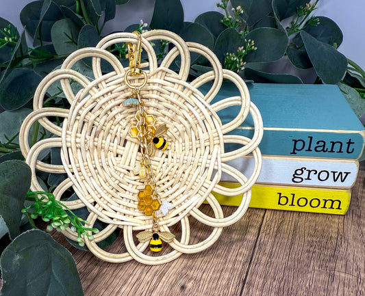 A woven basket with a bee charm and a stack of books with the words "plant grow bloom" written on them, all placed on a wooden surface with greenery in the background.