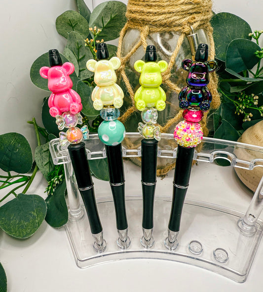 A clear plastic pen holder displaying four colorful pens with bear-shaped caps, placed on a white surface with greenery in the background.