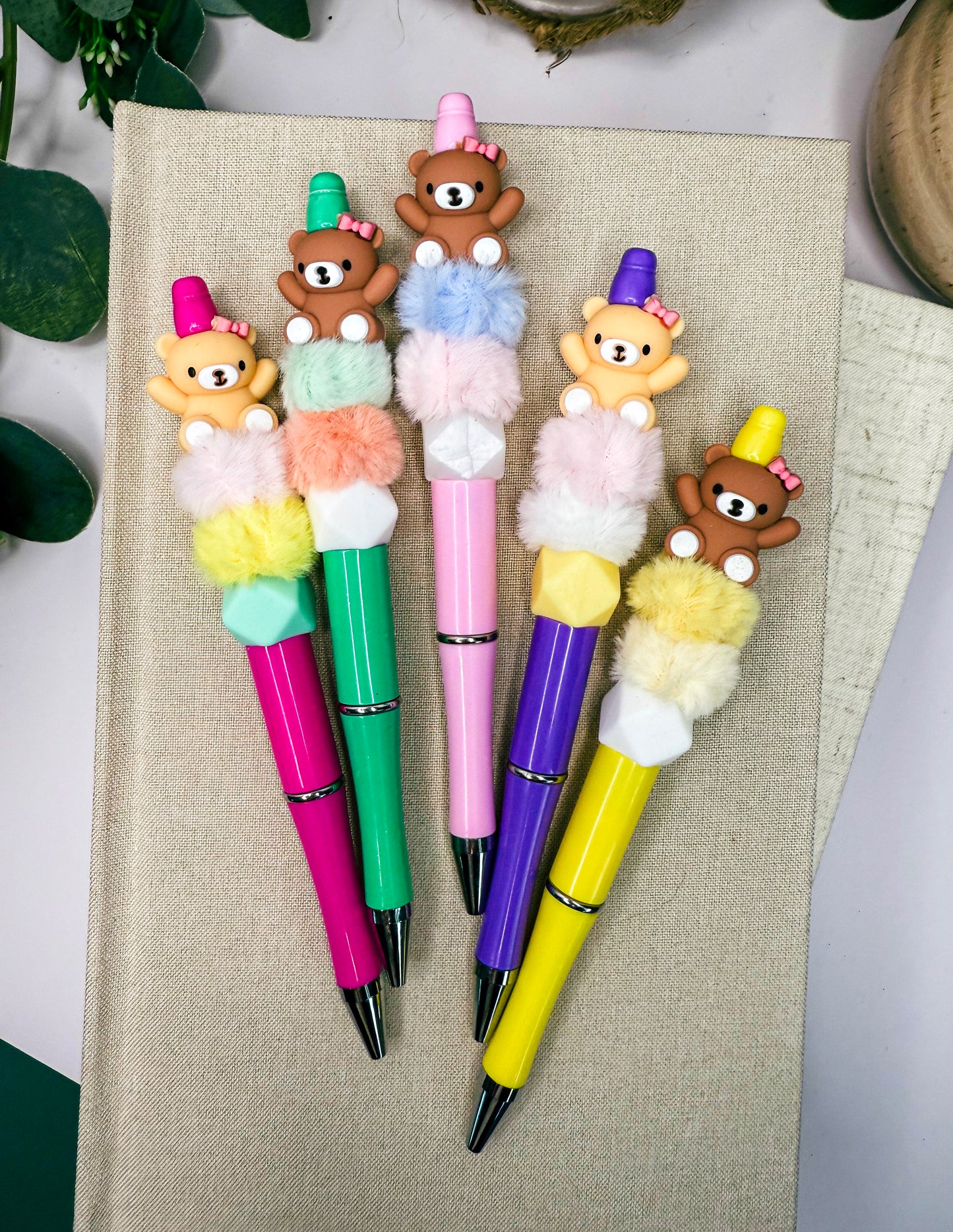 A collection of colorful pens with teddy bear designs on them, arranged on a beige surface.