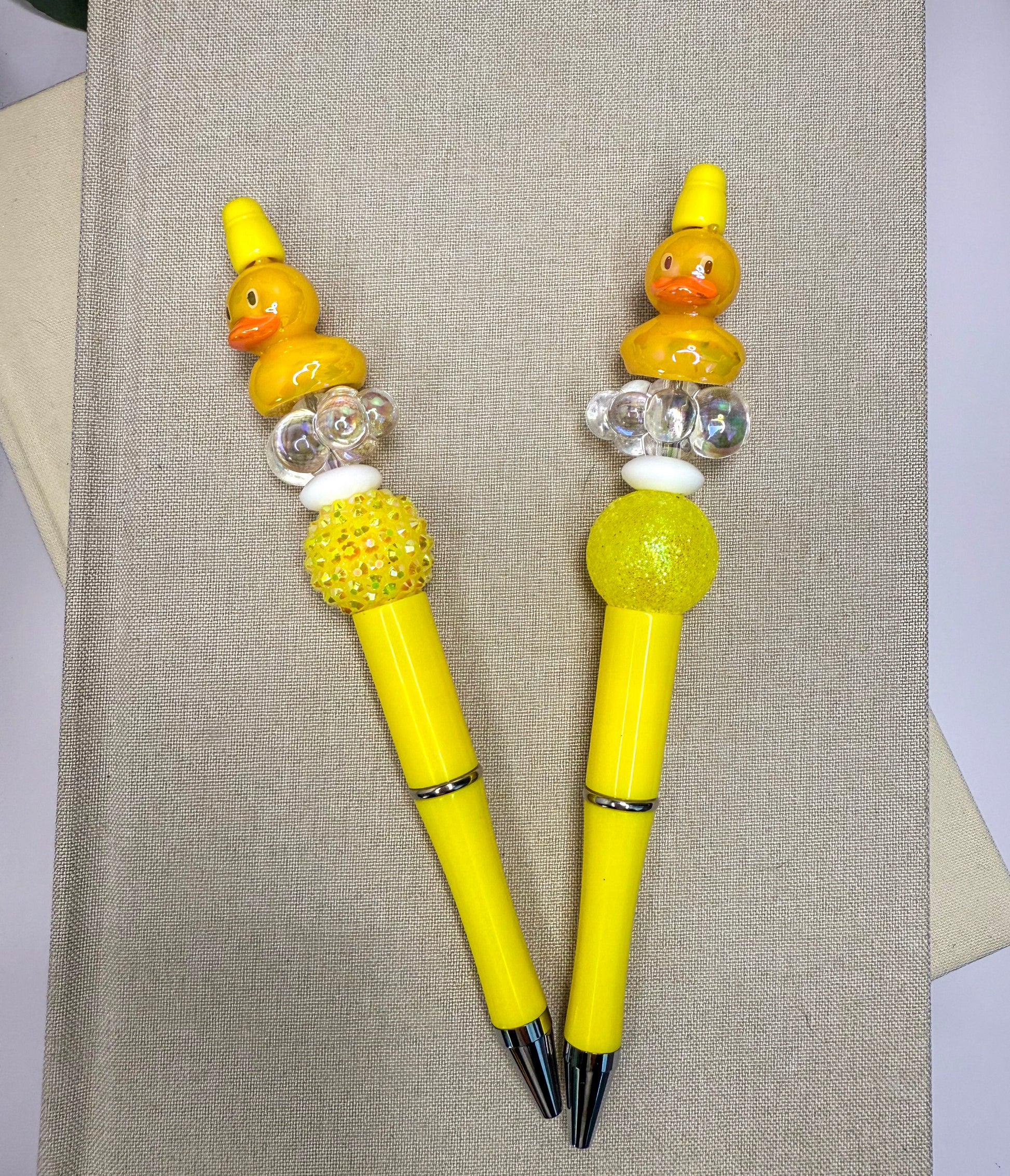 Two yellow pens with decorative elements, such as small rubber ducks and beads, are placed on a beige surface.