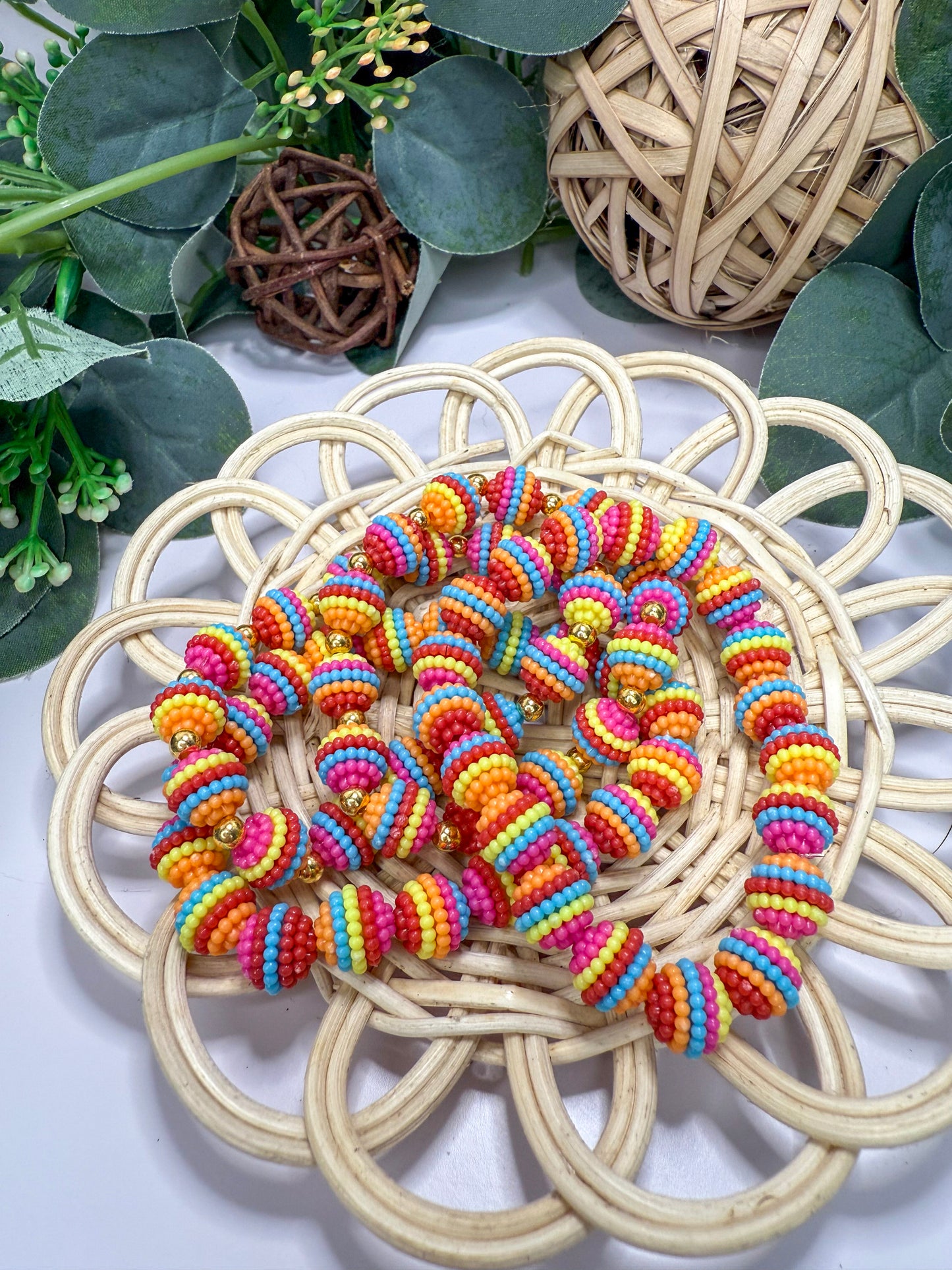 A colorful beaded necklace is displayed on a woven basket, surrounded by greenery and other natural elements.