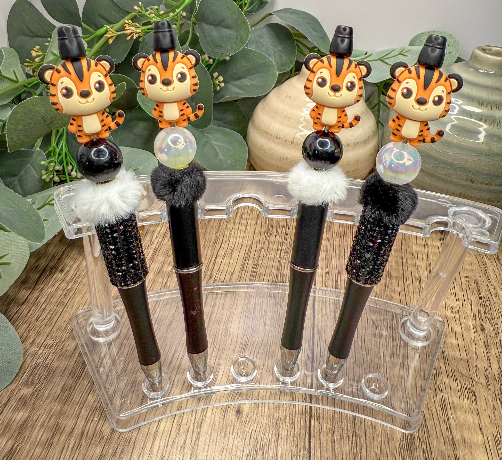 a collection of four novelty pens, each adorned with a tiger figurine on top. The pens are displayed in a clear plastic holder, which is placed on a wooden surface. The pens are arranged in a row, with each one featuring a unique design and color scheme. The tiger figurines on the pens add a playful and whimsical touch to the overall presentation.