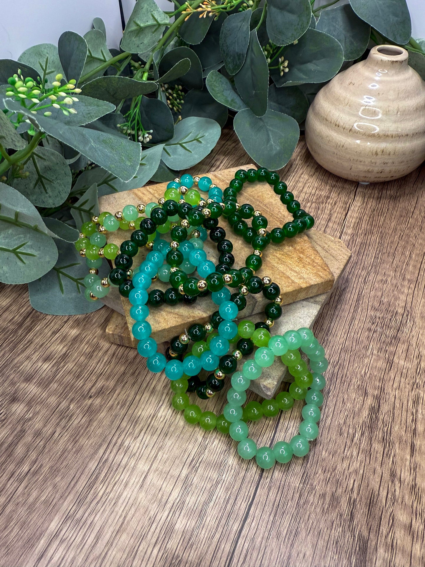 a collection of green and blue beaded bracelets arranged on a wooden surface, with a vase and greenery in the background.
