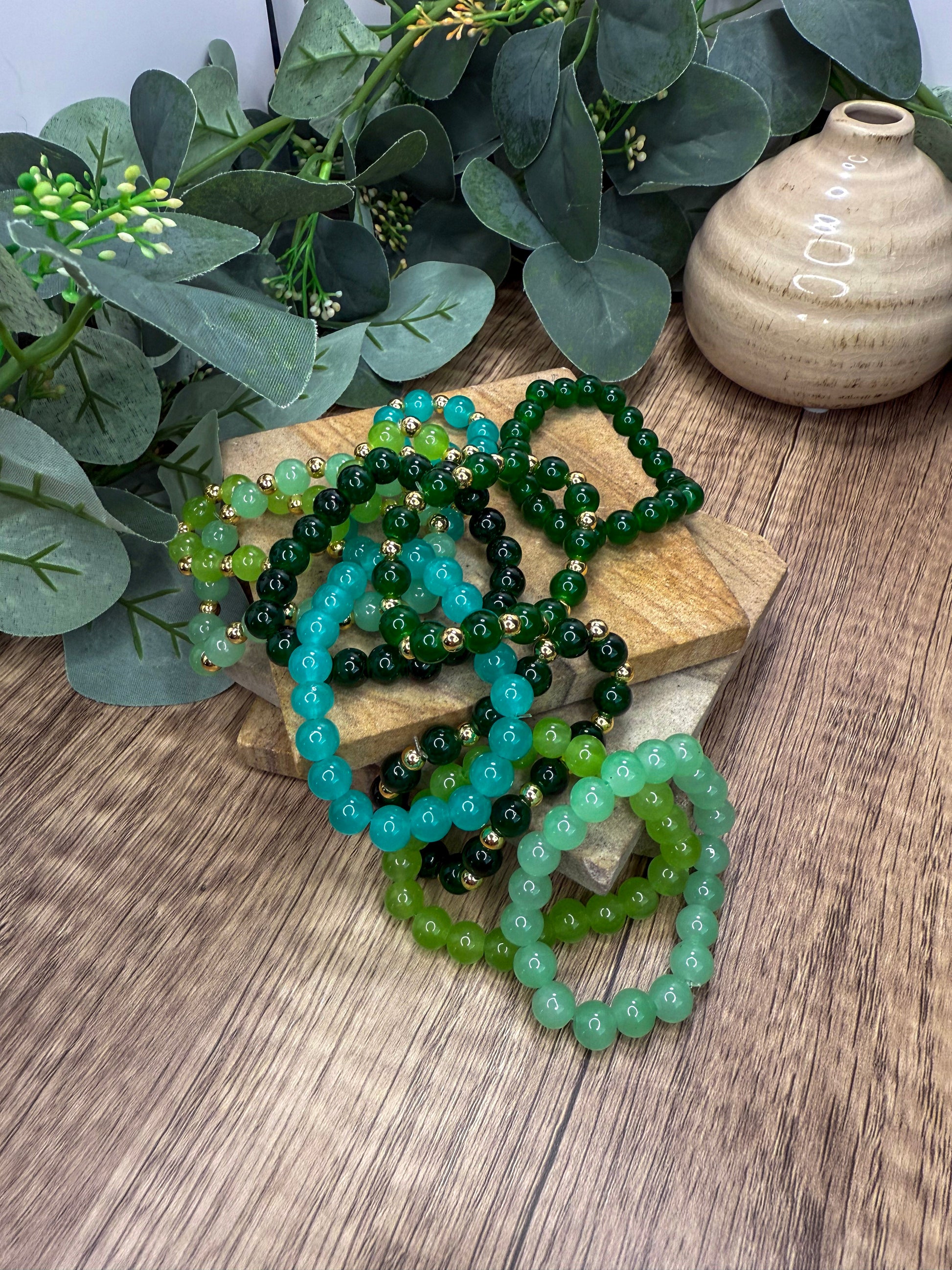 a collection of green and blue beaded bracelets arranged on a wooden surface, with a vase and greenery in the background.