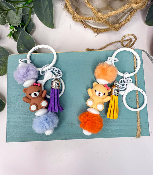 two keychains with tassels in the shape of teddy bears, one brown and one yellow, placed on a blue surface.