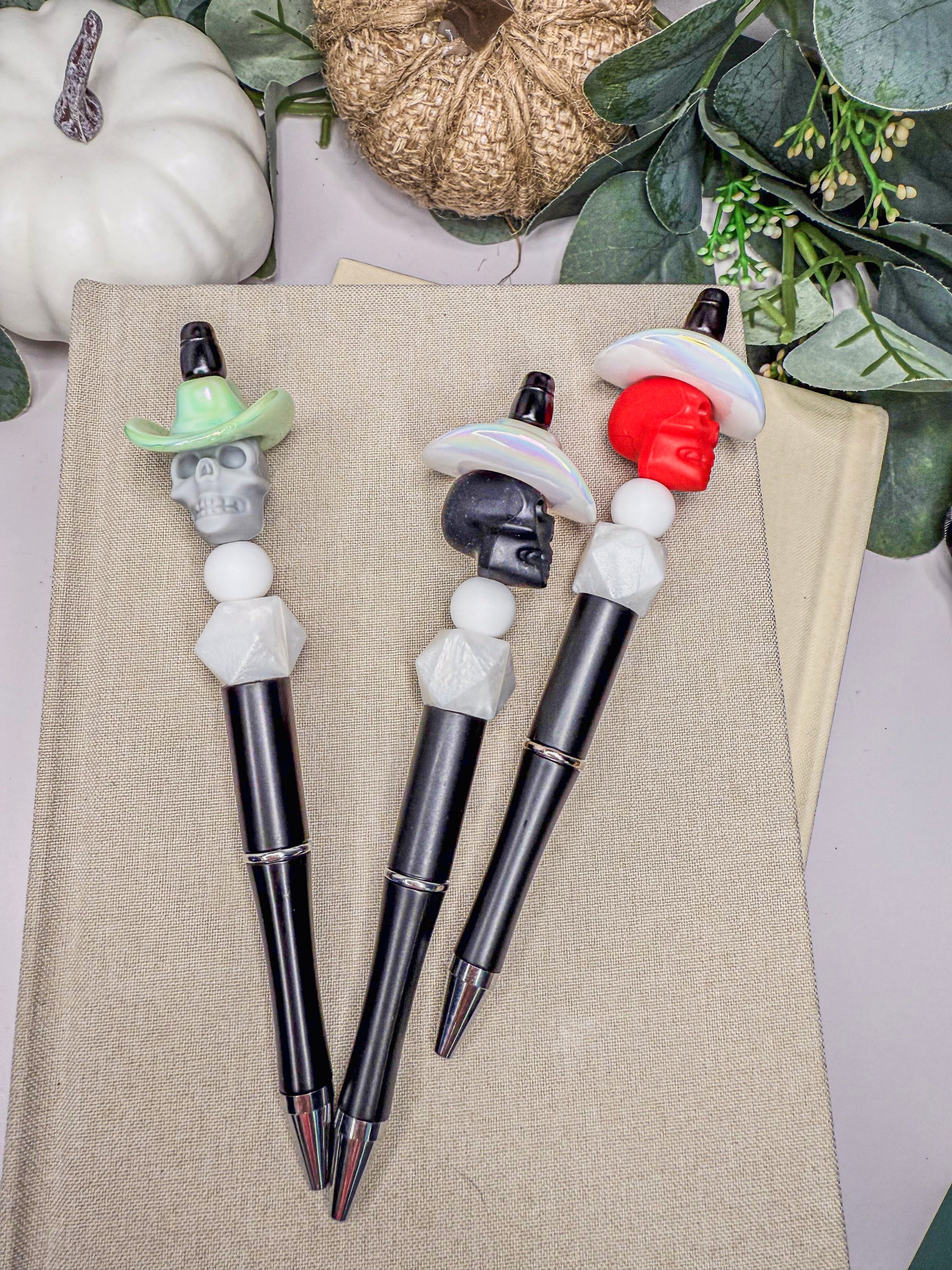 three pens with unique skull-shaped caps, arranged on a beige surface.