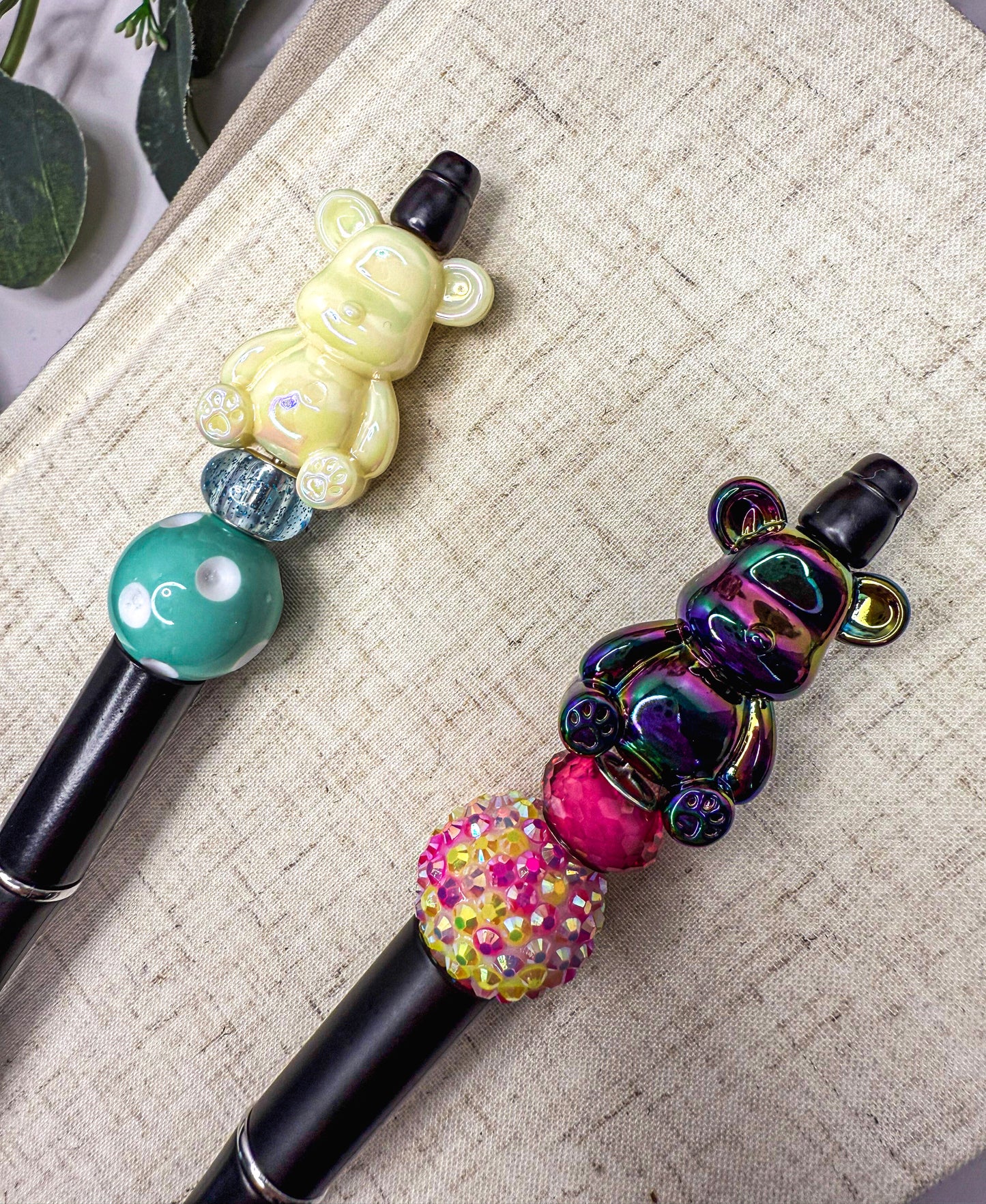two colorful pen-like objects with various decorations, including a yellow bear-shaped object and a purple bear-shaped object, placed on a textured surface.
