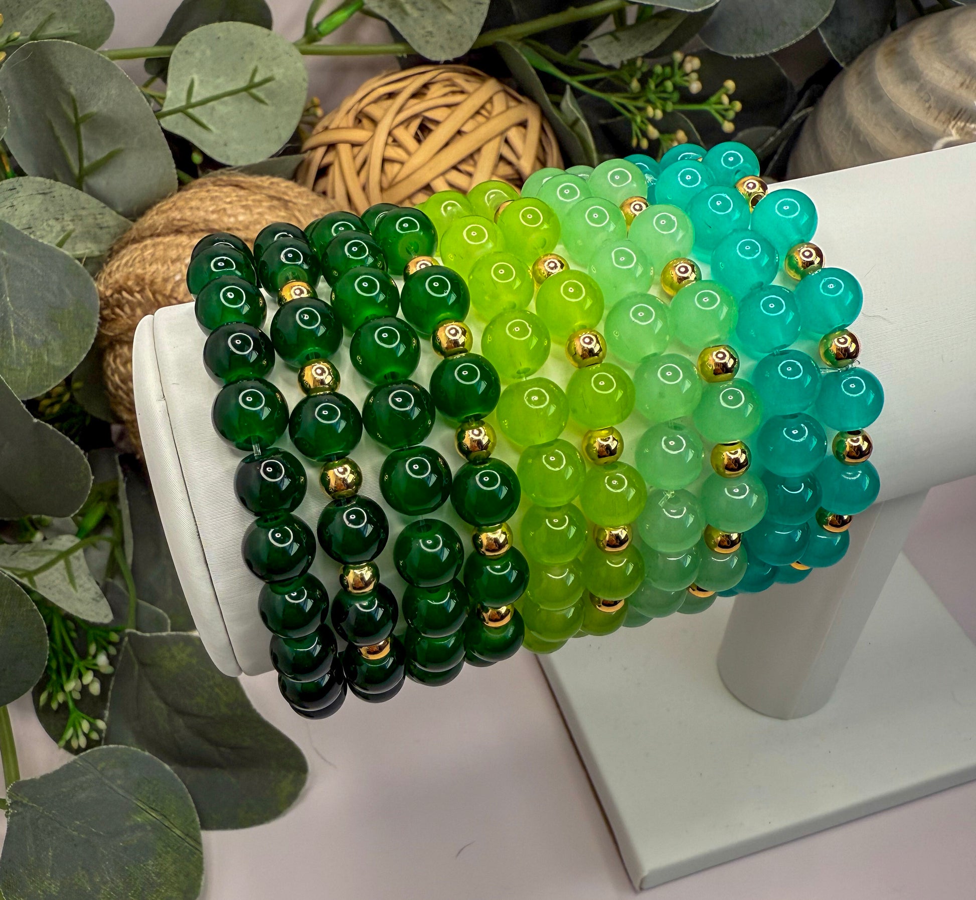 A white display stand holding a collection of colorful beaded bracelets in various shades of green and turquoise.