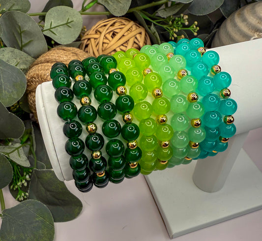 A white display stand holding a collection of colorful beaded bracelets in various shades of green and turquoise.