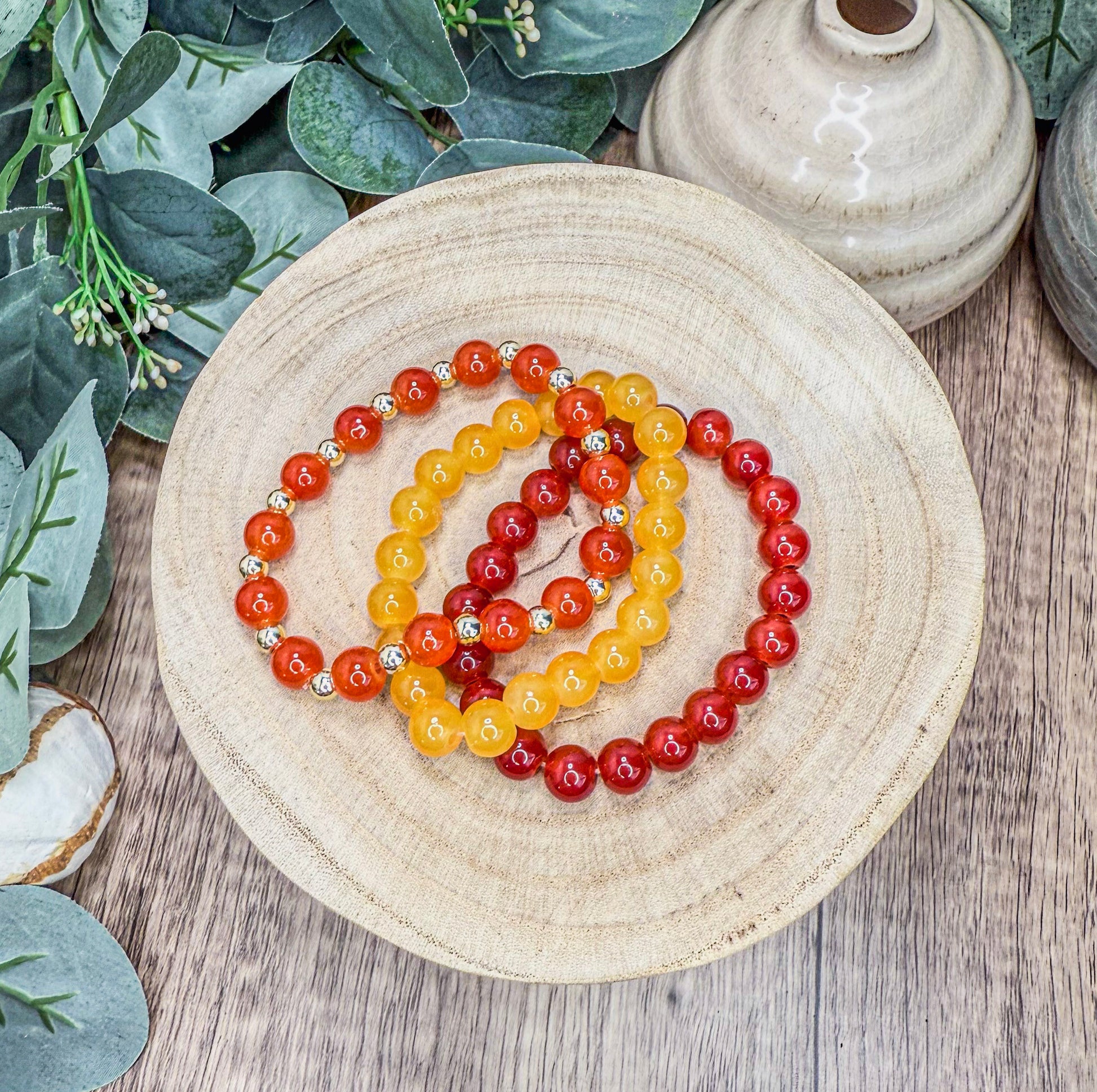 A wooden bowl holds a collection of colorful beaded bracelets, with a white vase and greenery in the background.