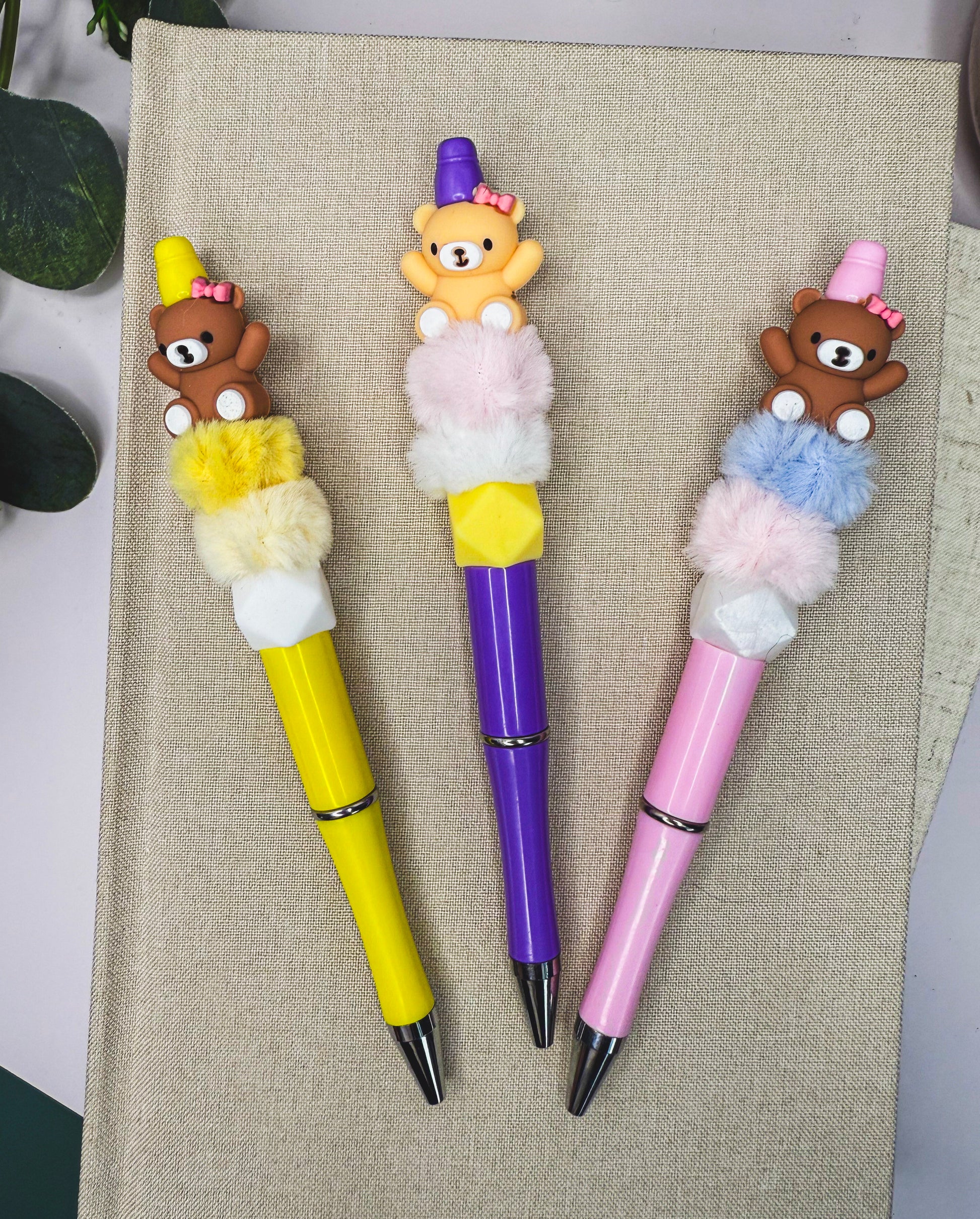 Three colorful pens with teddy bear decorations are placed on a beige surface.