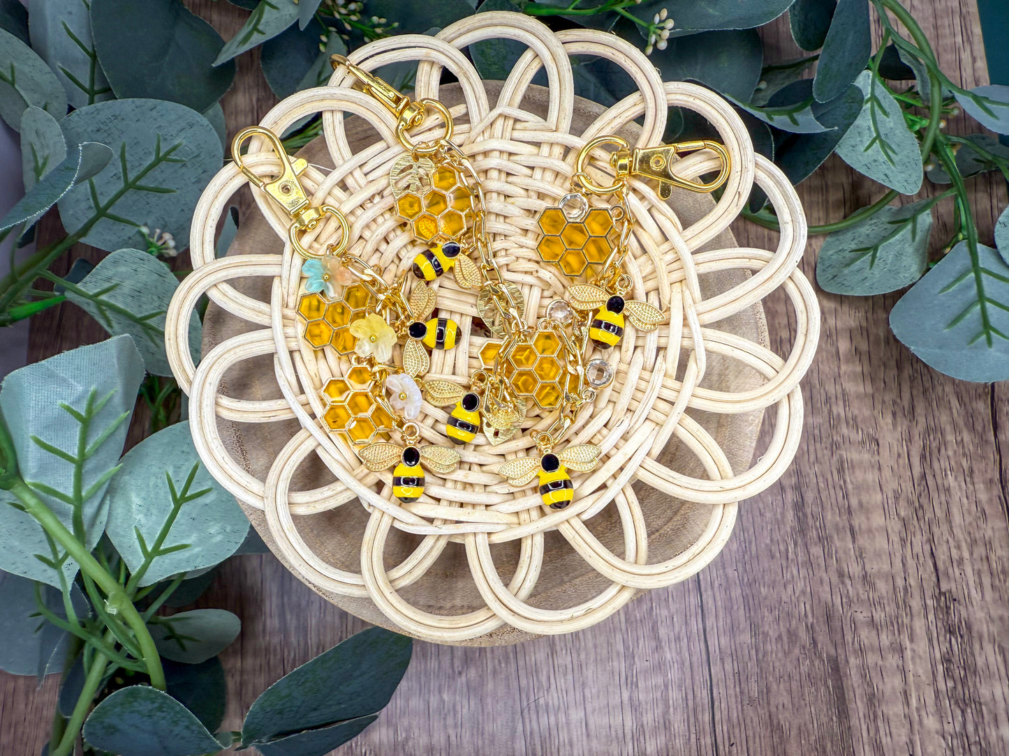 A pair of gold-colored earrings with honeycomb and bee-shaped charms are displayed on a woven basket, surrounded by green leaves.