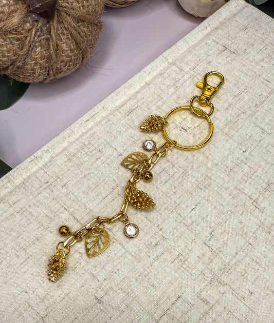 A gold-colored keychain with various charms, including pine cones and leaves, attached to a metal ring.