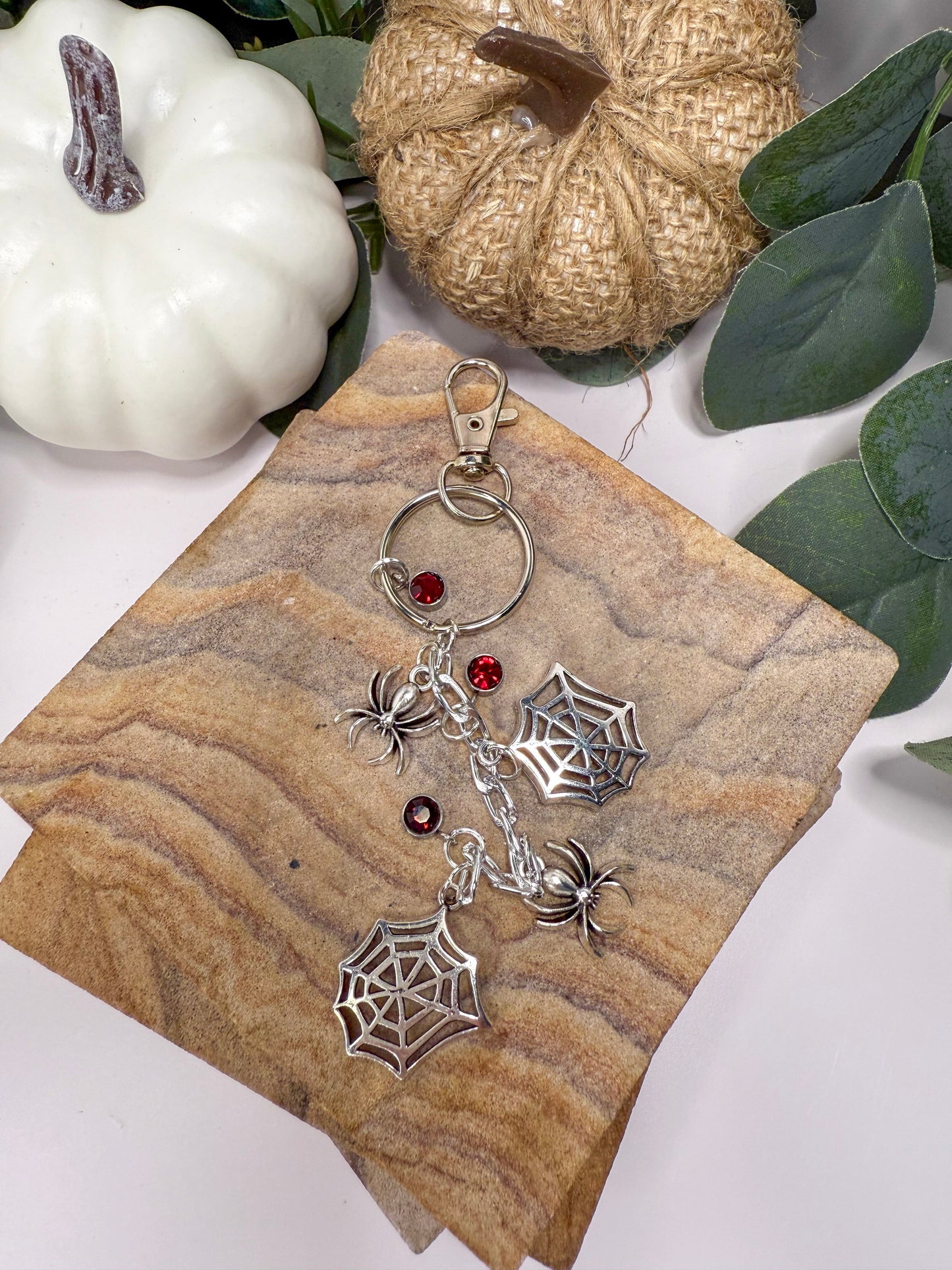 A keychain with a spider web design and red gemstones is displayed on a wooden surface, surrounded by small pumpkins and greenery.