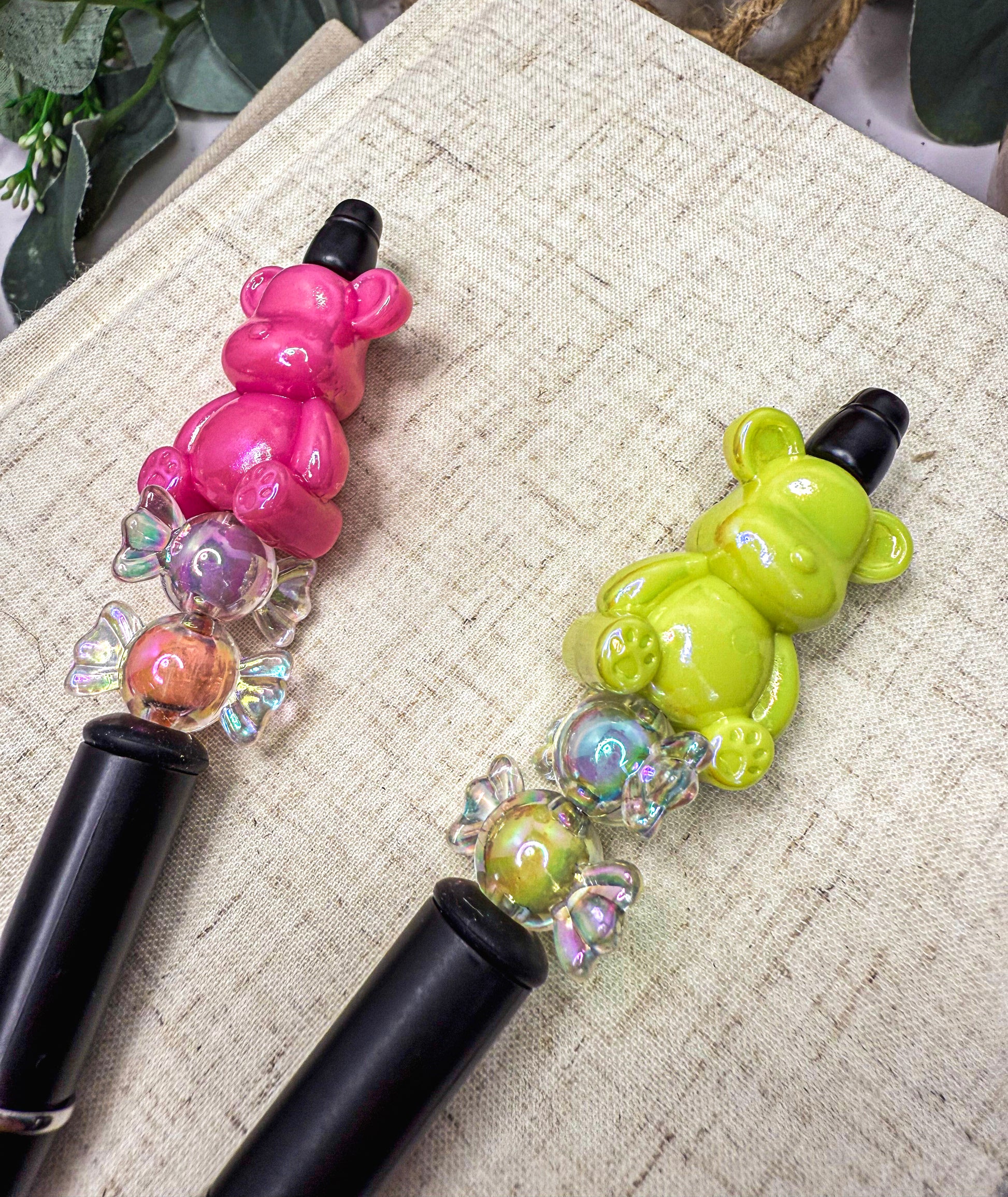 Two colorful pen-like objects with bear-shaped heads are placed on a textured surface.