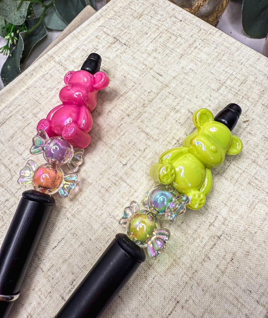 Two colorful pen-like objects with bear-shaped heads are placed on a textured surface.