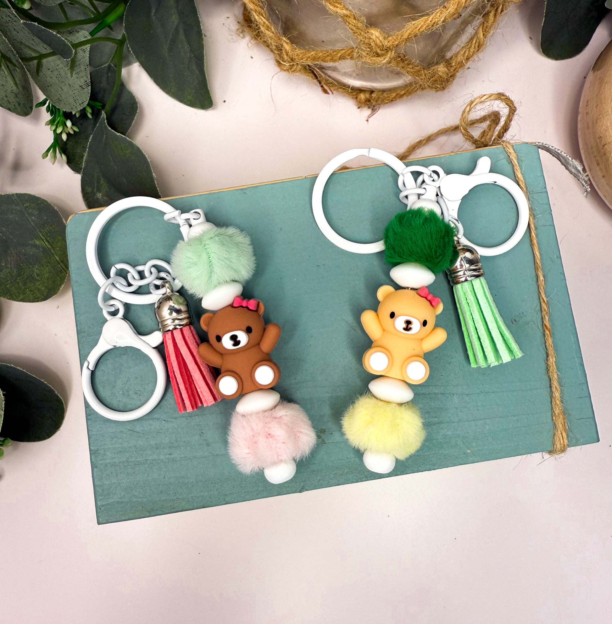 two keychains with tassels and small teddy bear figures attached to them, placed on a blue surface.