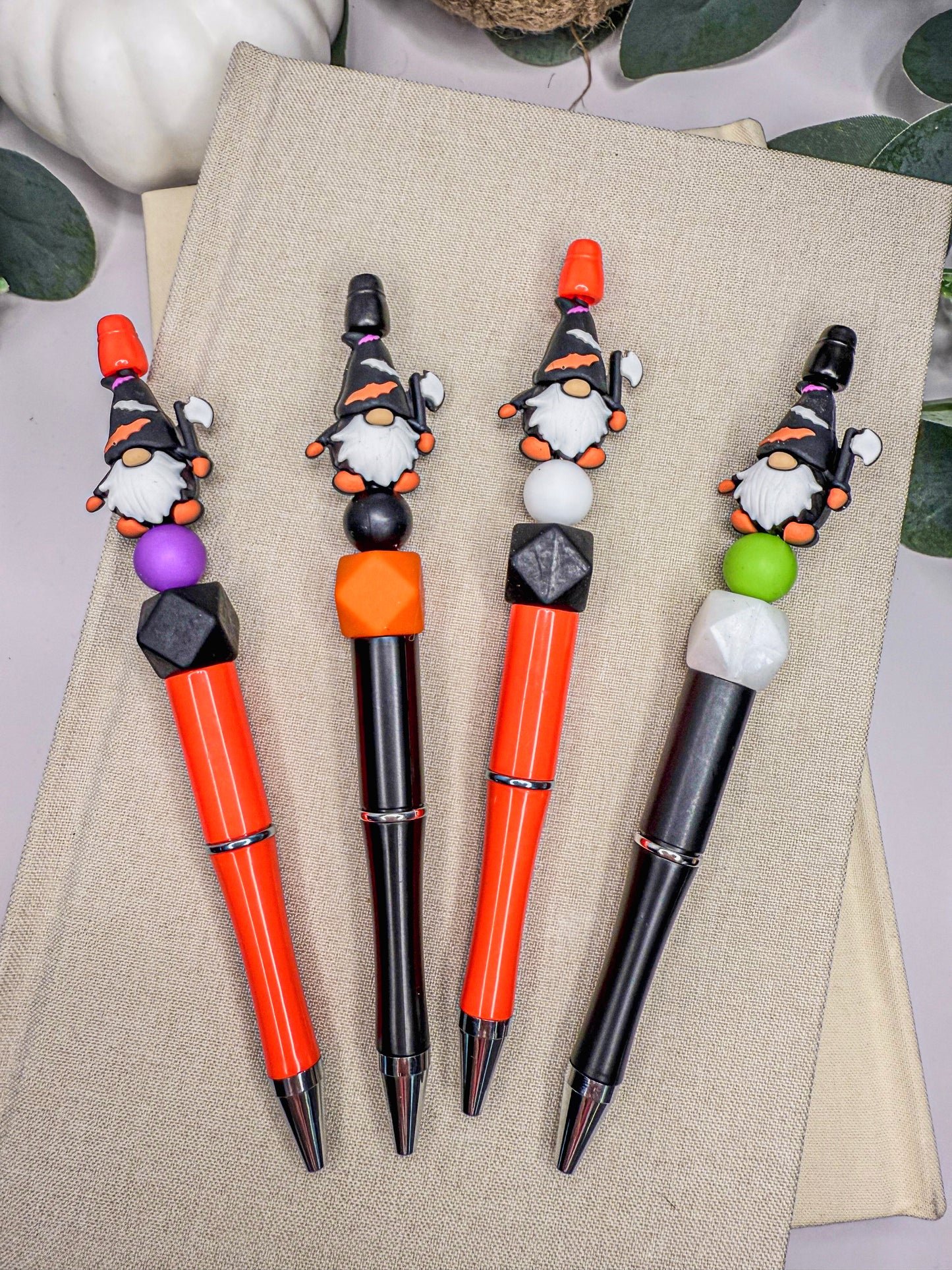 four pens with unique designs, including gnome-like figures and colorful beads, arranged on a beige surface.