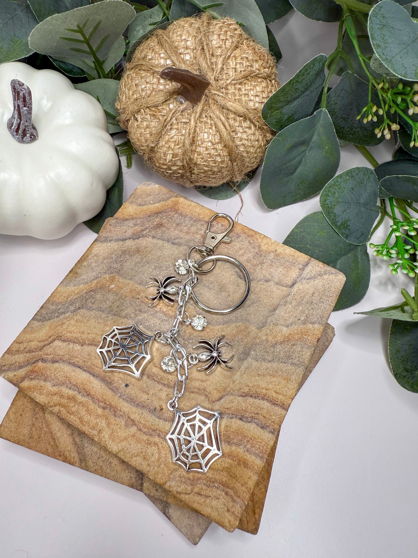 a keychain with a spider web design, placed on a wooden surface alongside other decorative items such as a pumpkin and greenery.