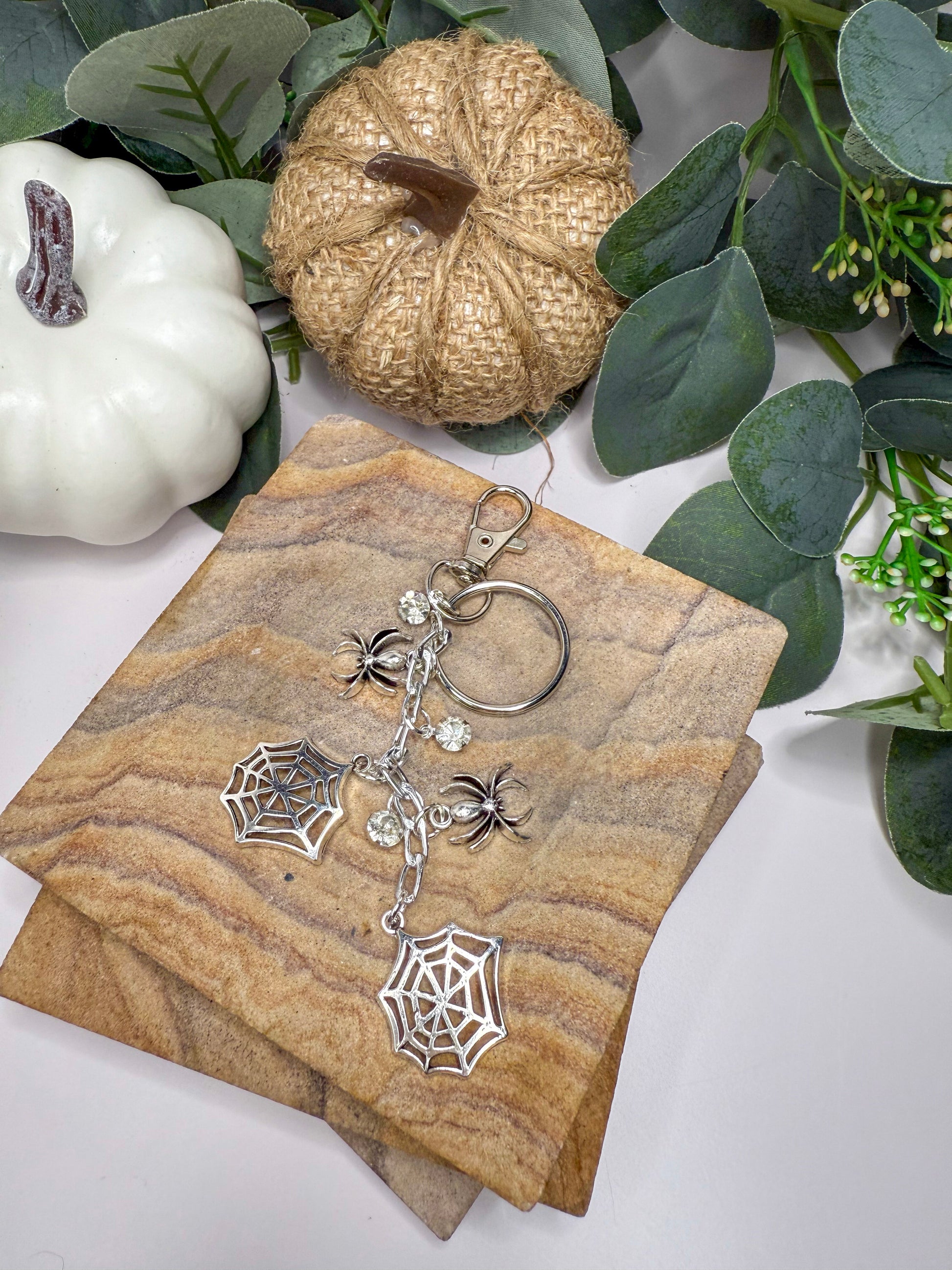 a keychain with a spider web design, placed on a wooden surface alongside other decorative items such as a pumpkin and greenery.