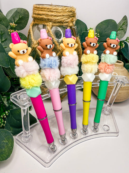 A display of five colorful pens with teddy bear decorations, arranged on a clear stand.