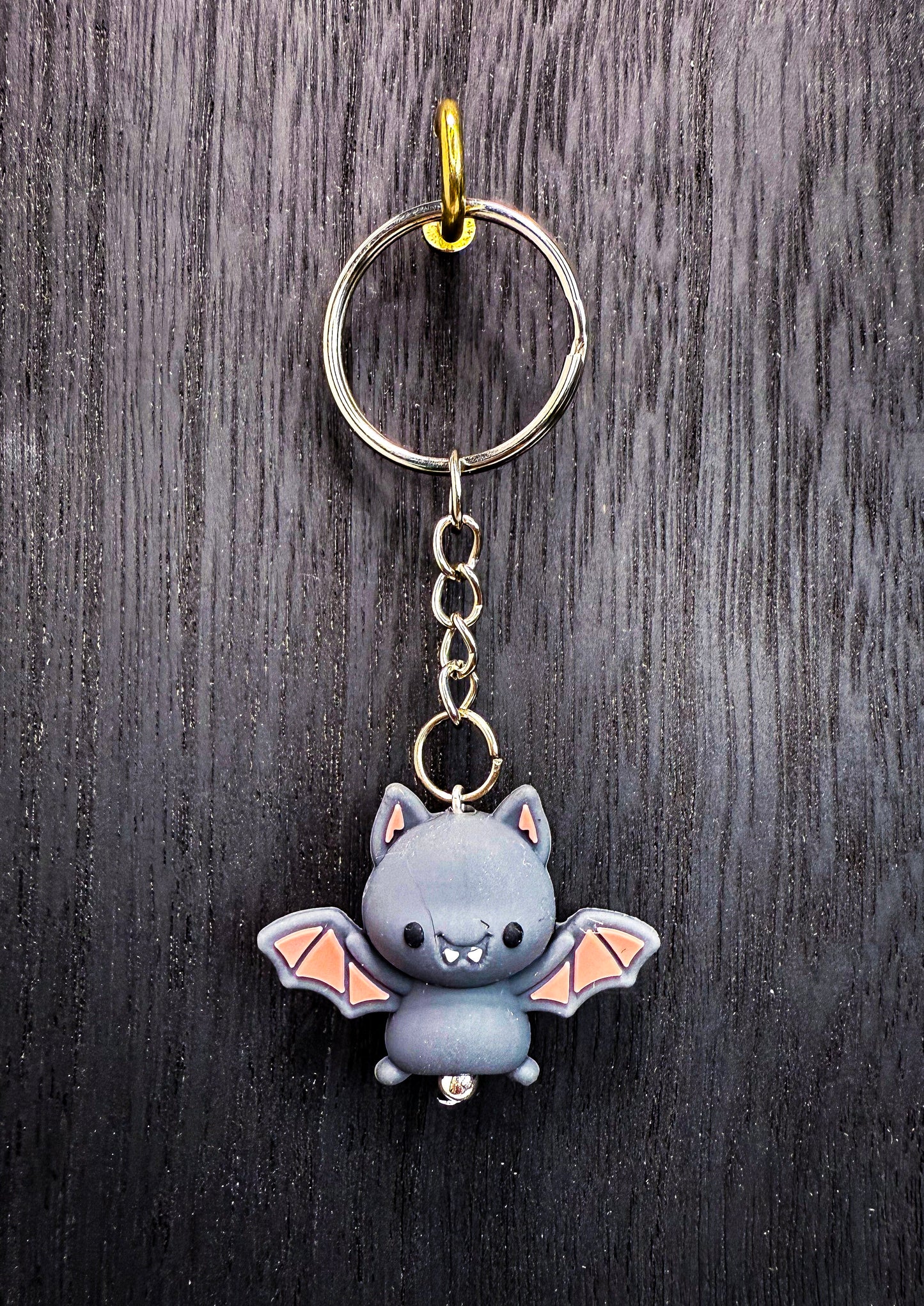 A keychain featuring a cute gray bat with orange wings, hanging on a hook against a dark wooden background.