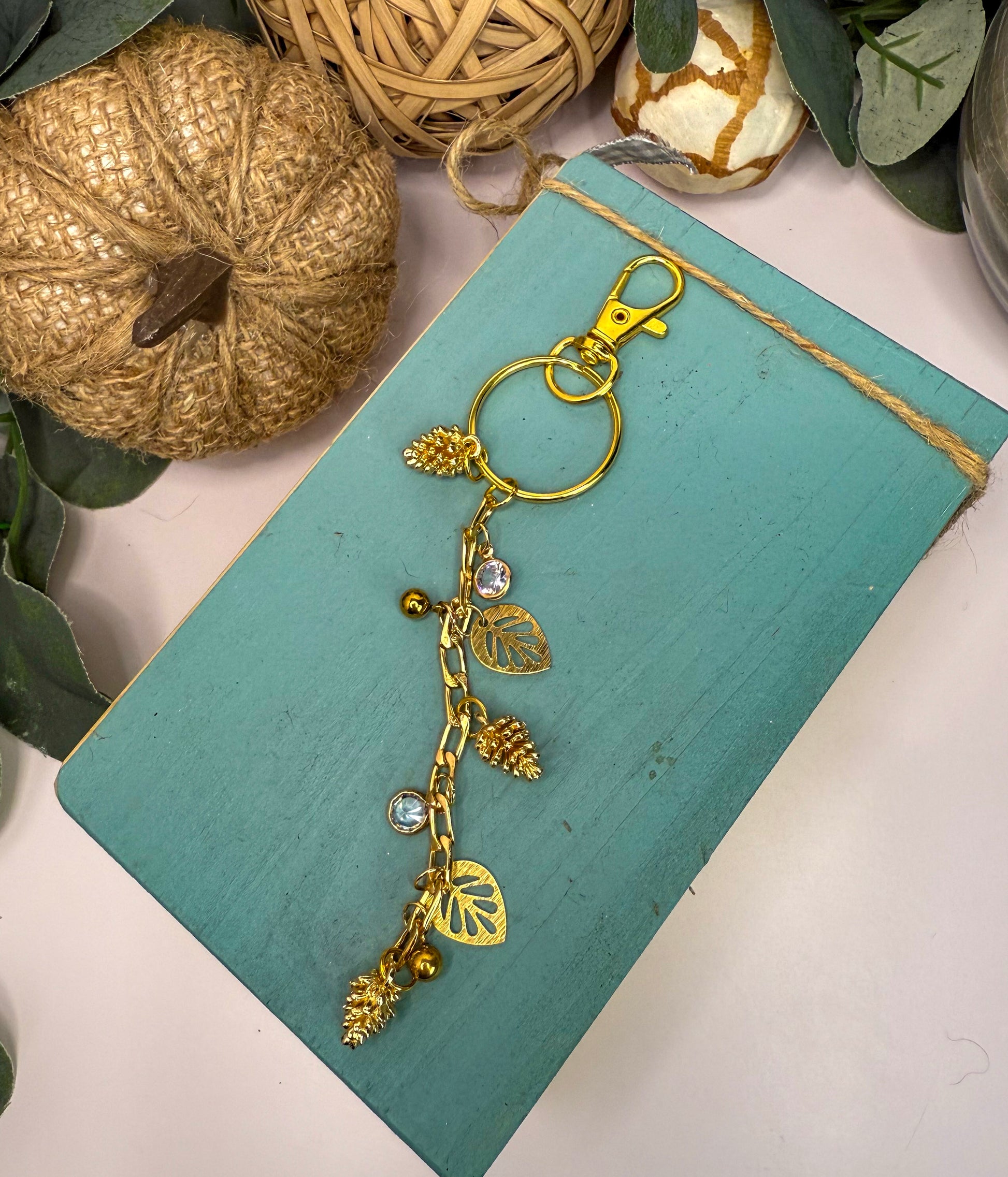 a turquoise-colored wooden surface with a gold-colored keychain hanging from it. The keychain has a leaf design and pine cone-shaped charms, giving it a unique and decorative appearance. The keychain is placed on a light-colored surface, and there are some decorative elements such as a pumpkin and a woven basket in the background.
