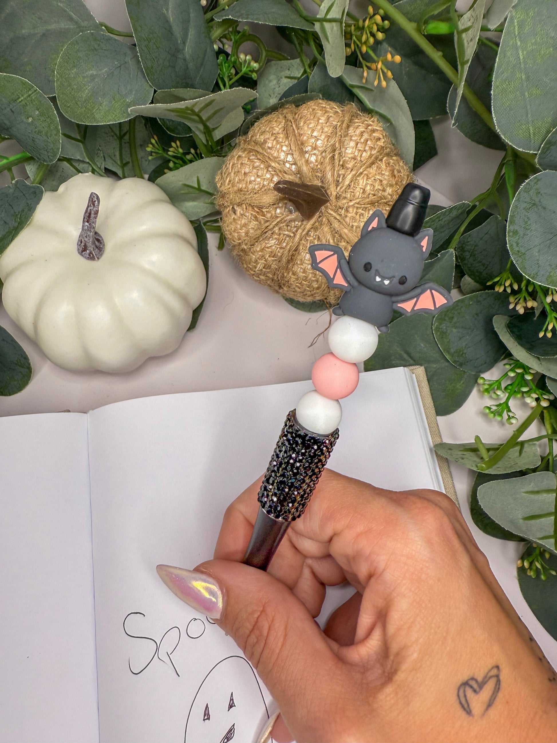 A hand holding a pen with a bat design, surrounded by Halloween decorations such as a pumpkin and a small stuffed animal.