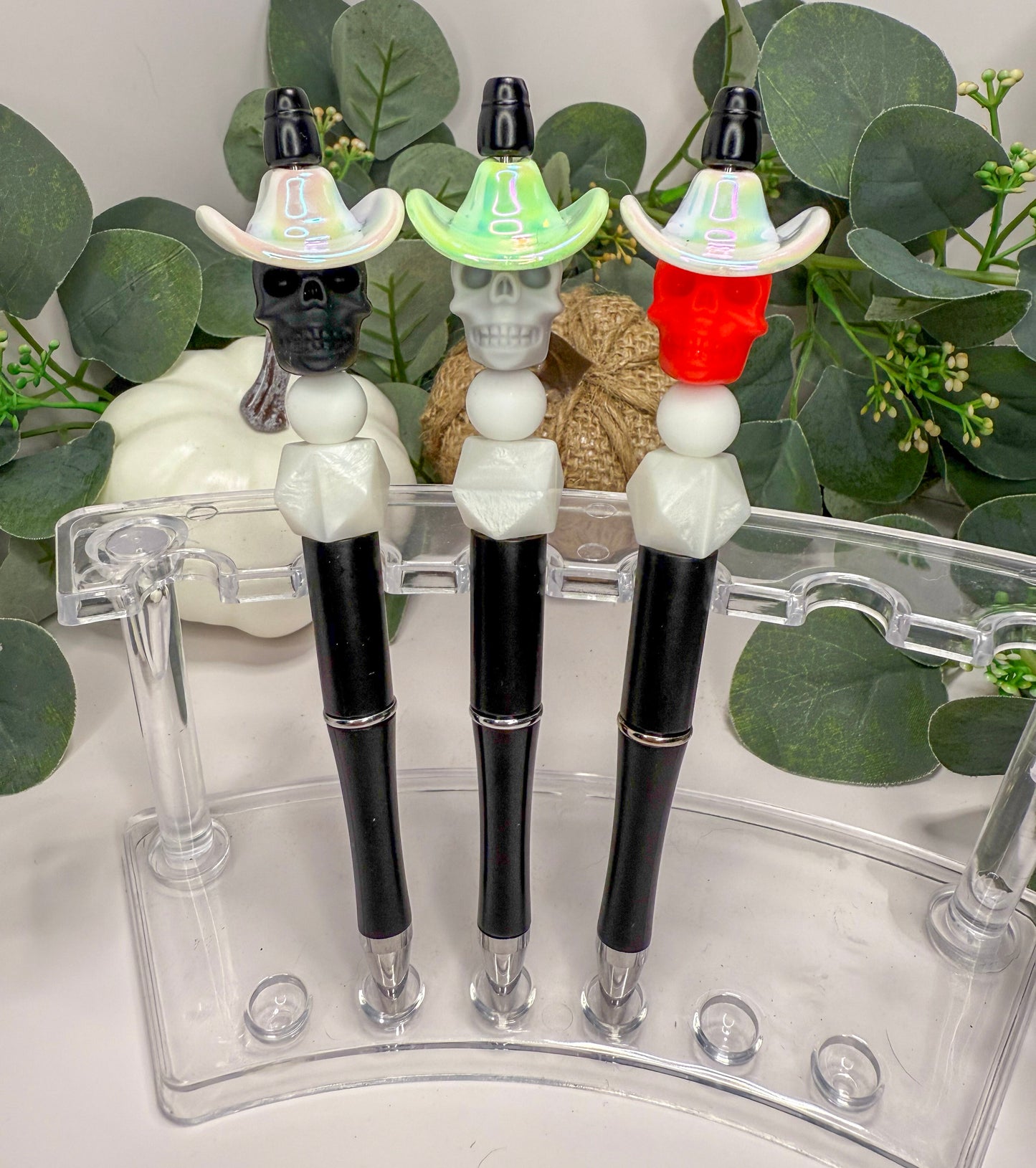 Three colorful pens with skull-shaped caps are displayed on a clear stand, surrounded by greenery.