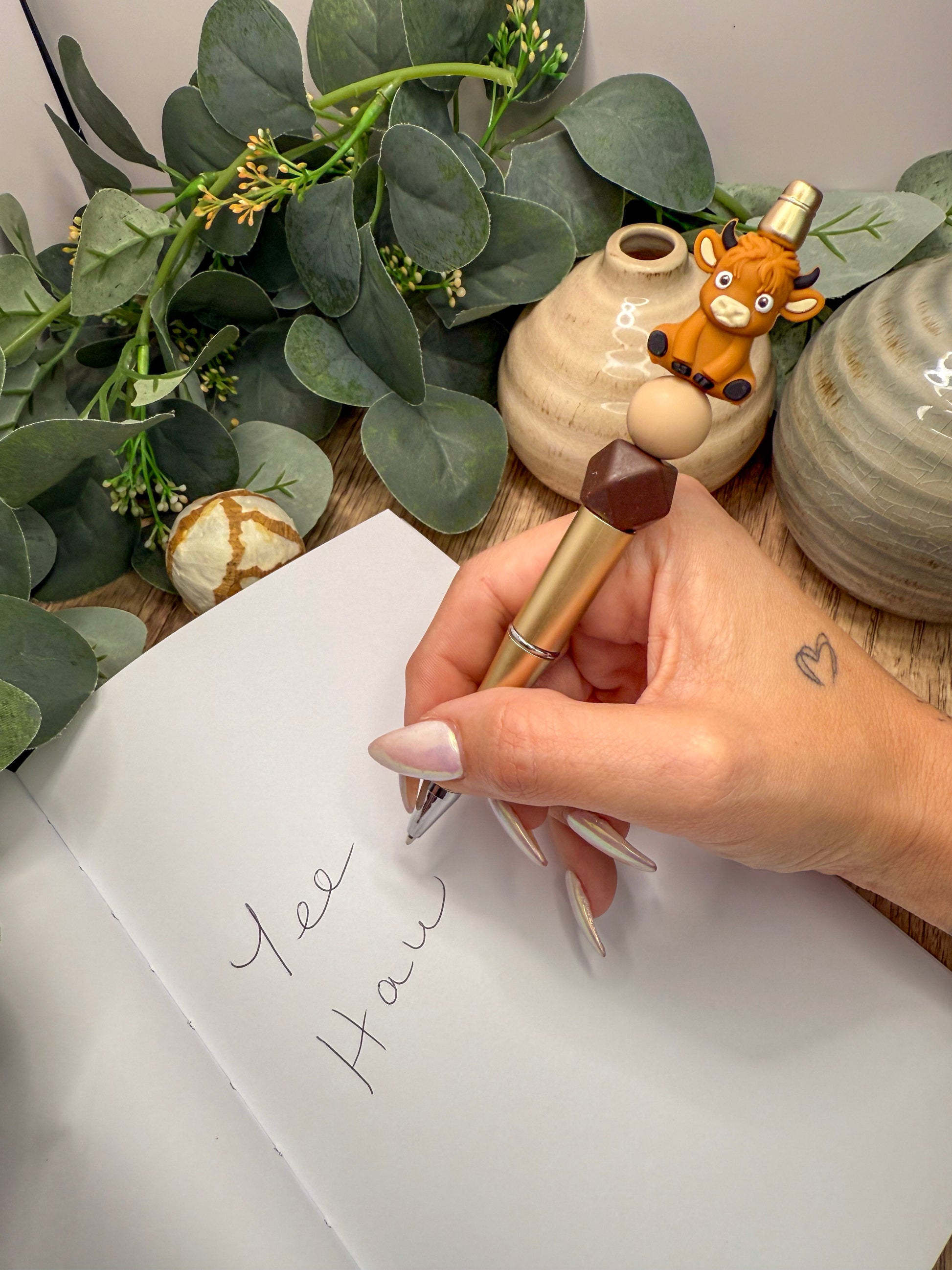 A hand holding a pen and writing on a notebook, with a small figurine of a cow and a plant in the background.