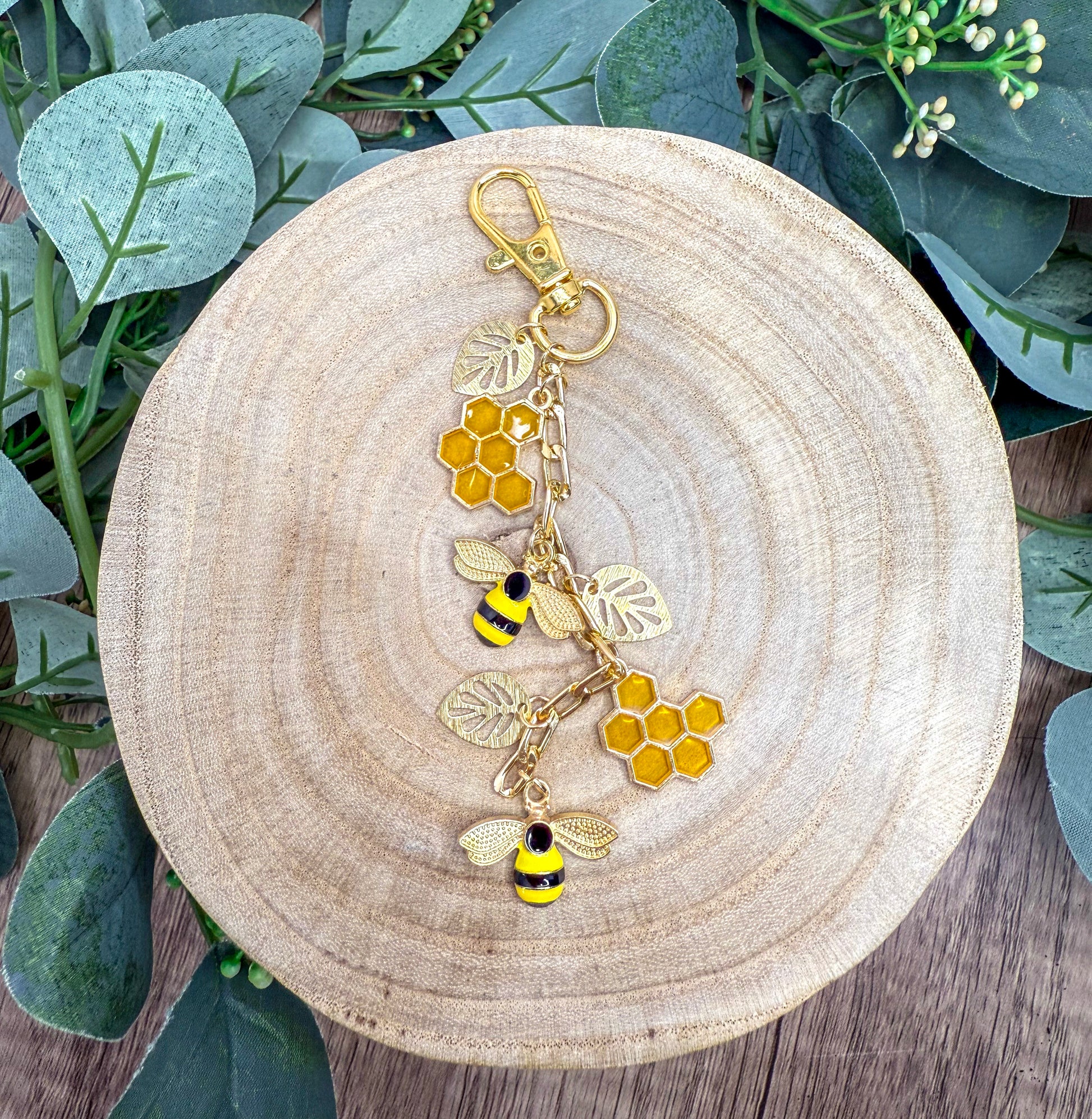 A wooden plate with a decorative keychain featuring a bee and honeycomb design, surrounded by greenery.