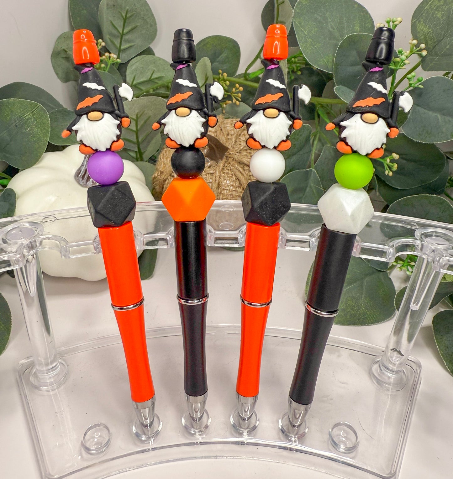 A collection of four colorful pens with whimsical gnome-like designs on the caps, arranged in a row on a clear stand.