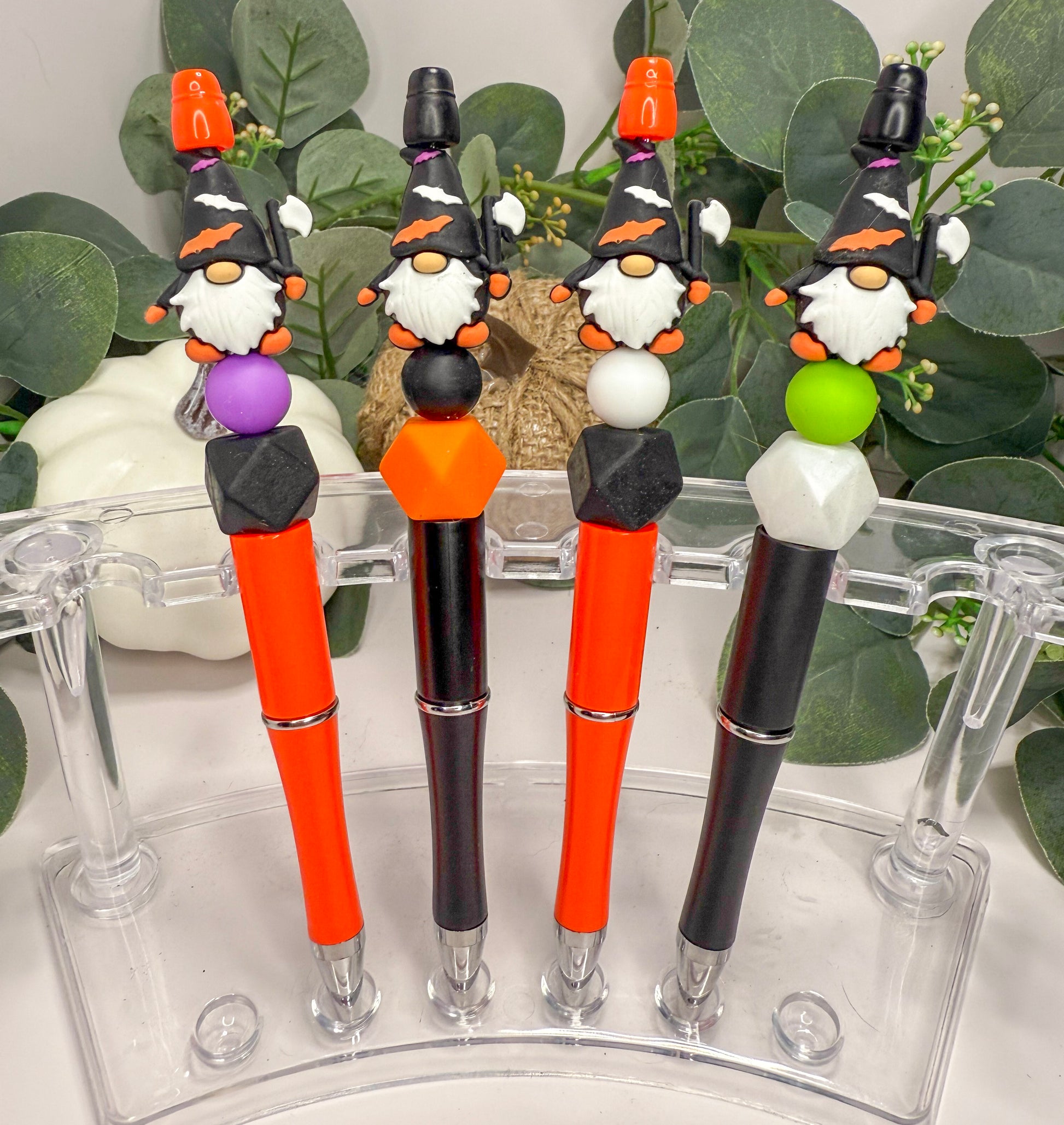 A collection of four colorful pens with whimsical gnome-like designs on the caps, arranged in a row on a clear stand.