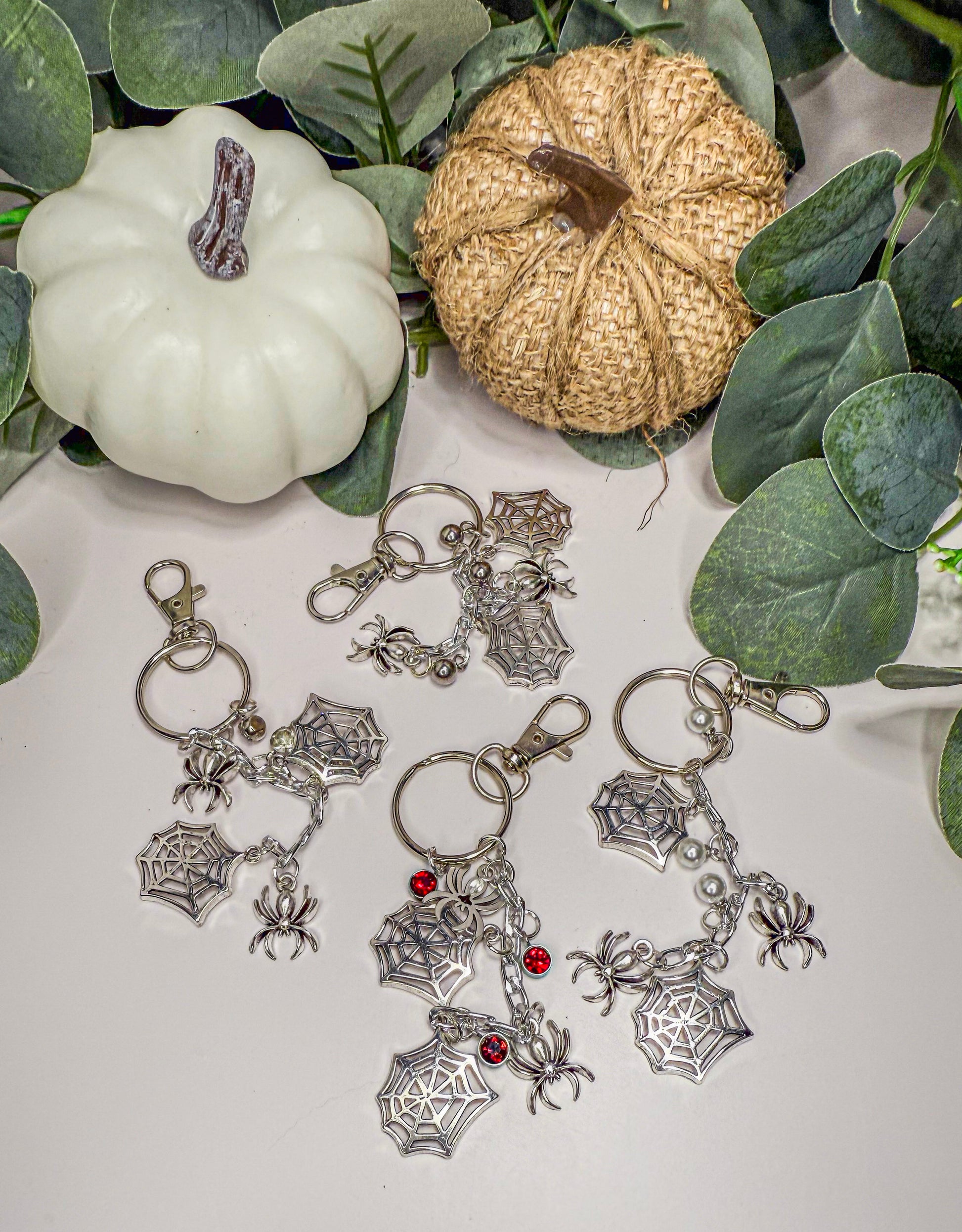 a collection of keychains with spider web charms, including red jewels, placed on a white surface. There are also two small pumpkins and some greenery in the background.