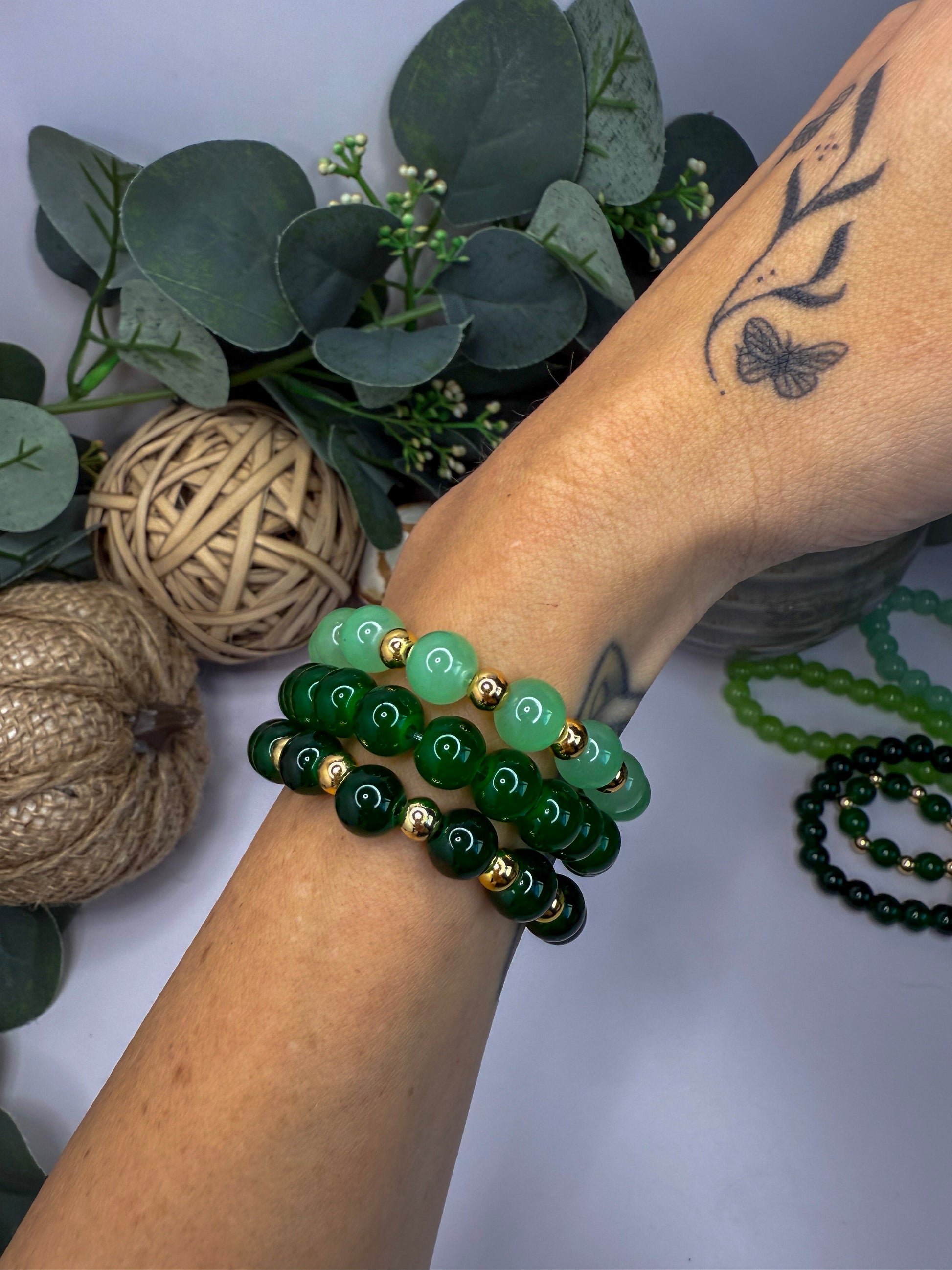 A person's arm wearing multiple green beaded bracelets, with a tattoo visible on their skin. The background includes some greenery and other decorative items.