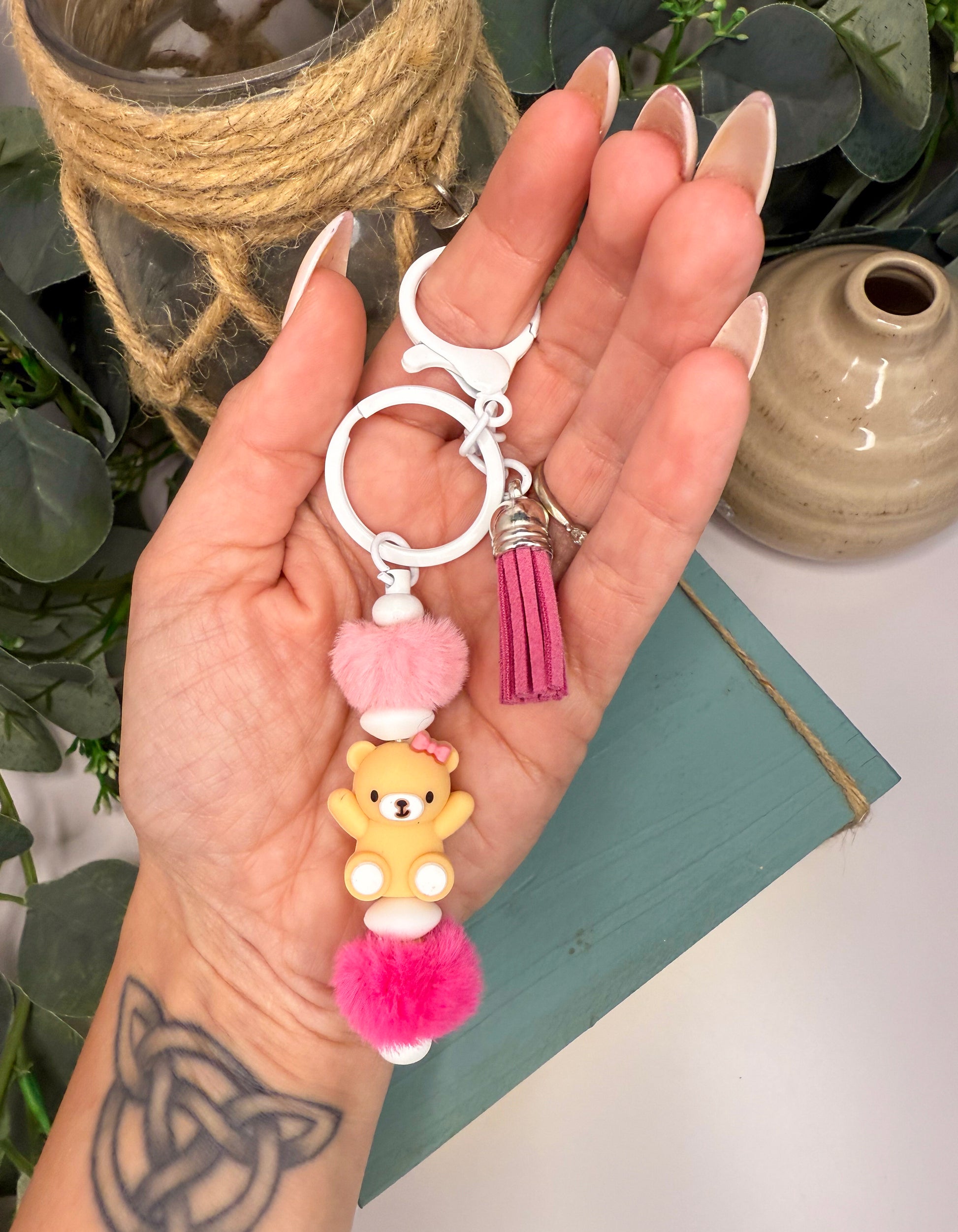 A hand holding a keychain with a small teddy bear charm and a pink tassel.
