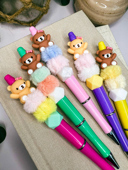 A collection of colorful pens with teddy bear decorations, including brown, yellow, pink, green, and purple designs.