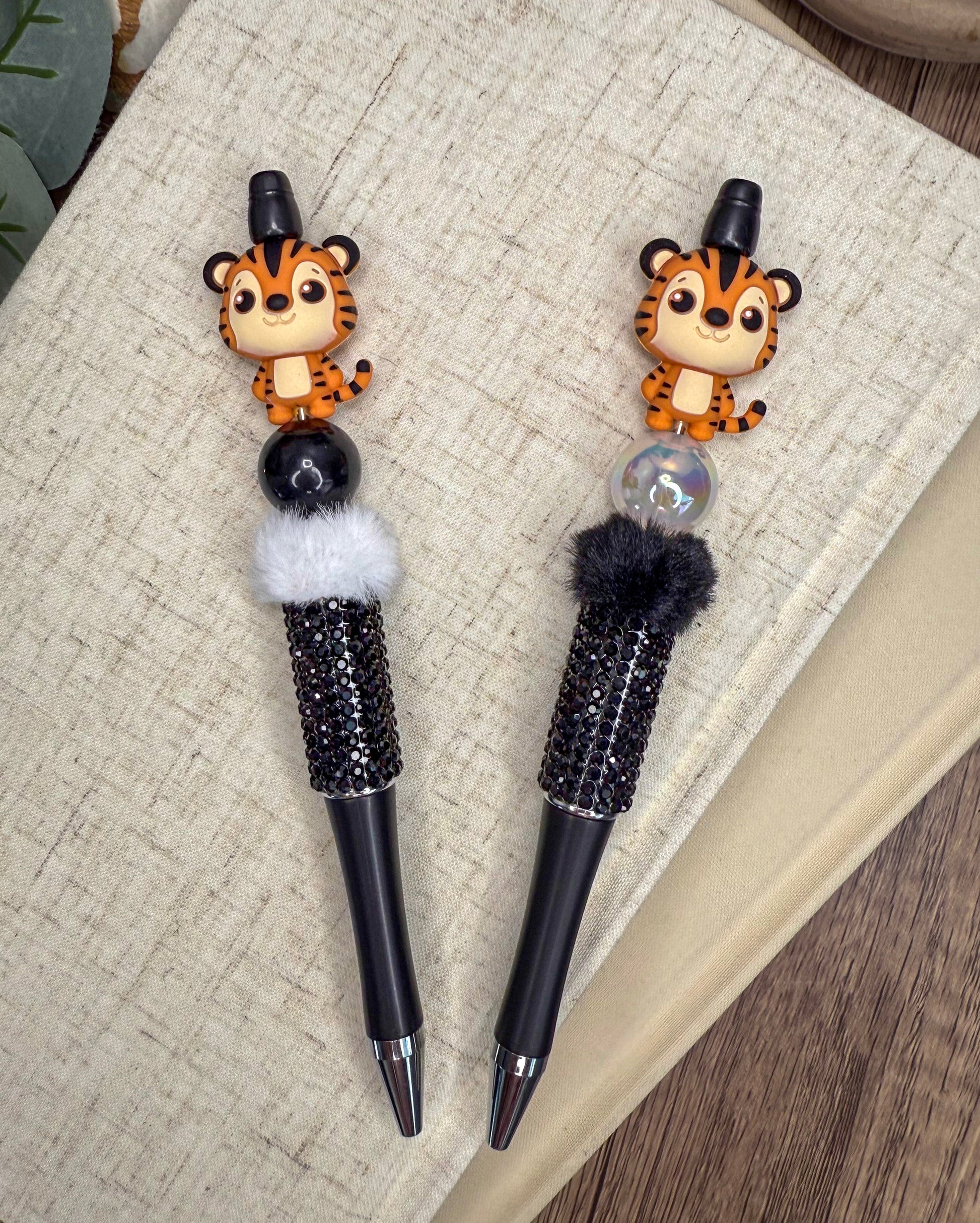 two pens with tiger figurines on the caps, placed on a textured surface.