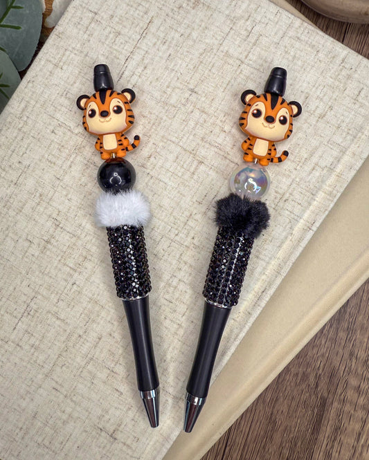 two pens with tiger figurines on the caps, placed on a textured surface.