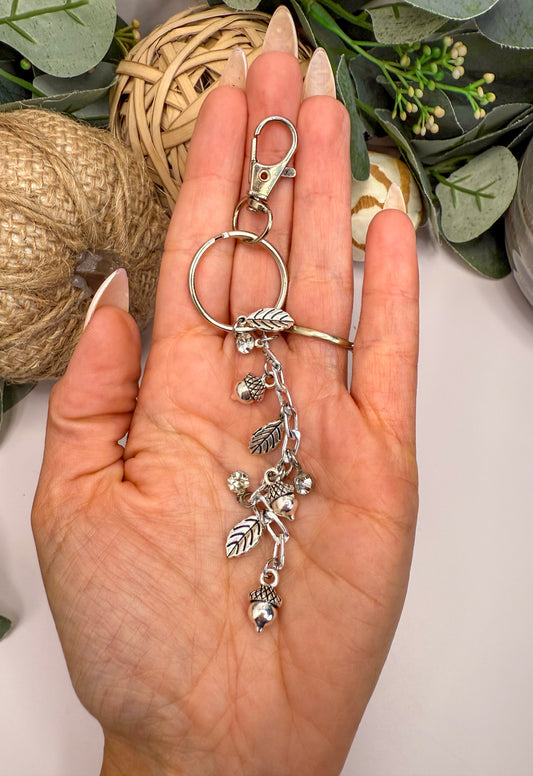A hand holding a silver keychain with various charms, including leaves and acorns, against a backdrop of greenery.