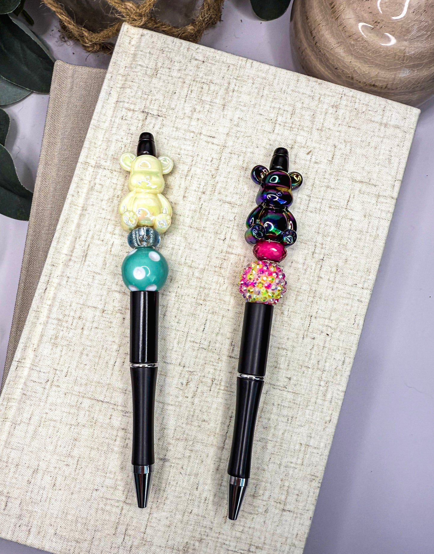 Two colorful pens with bear-shaped charms are placed on a beige surface, with a small wooden object and some greenery in the background.