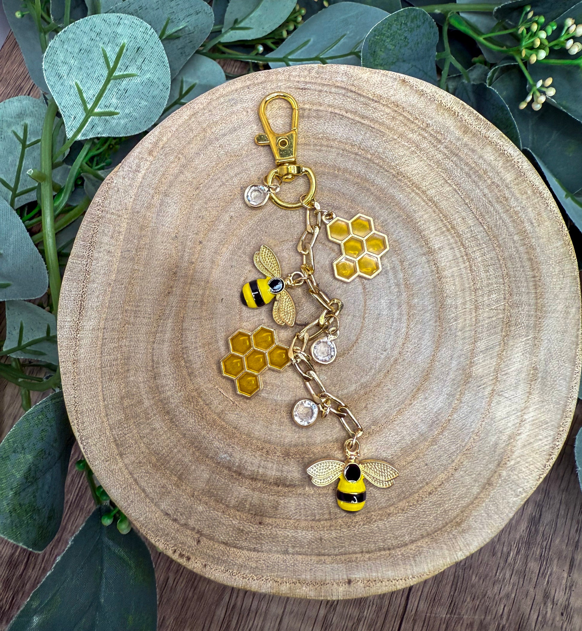 A wooden plate with a decorative keychain featuring a bee and honeycomb design, surrounded by greenery.