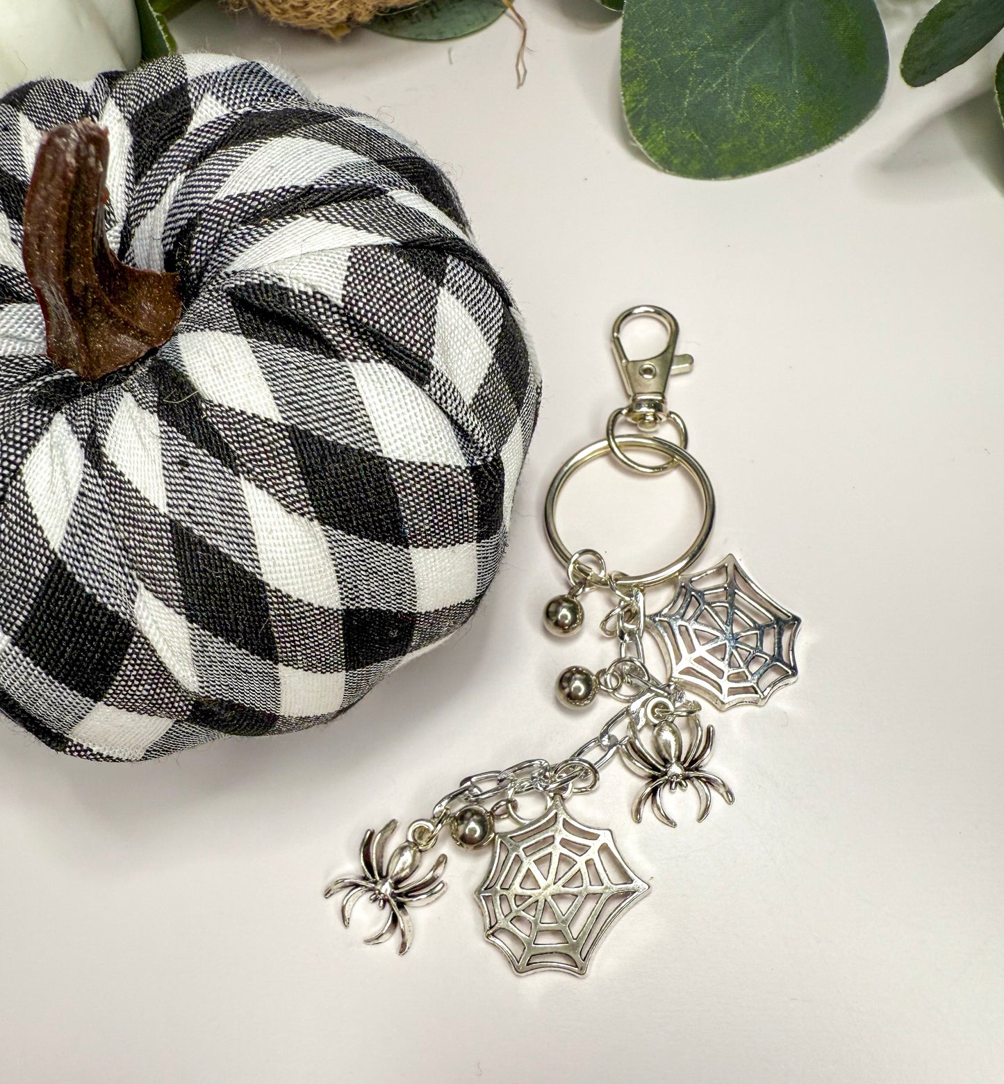 a black and white checkered pumpkin and a silver keychain with spider charm and bell charms, placed on a white surface.