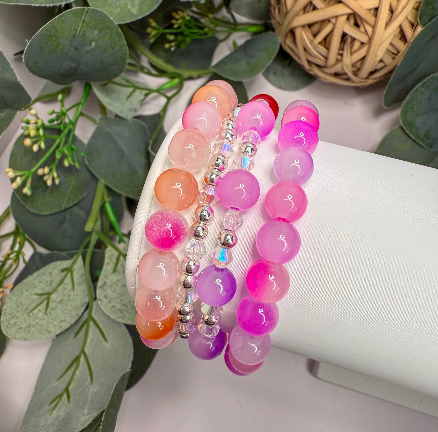 A white bracelet display with three beaded bracelets in various colors, including pink, purple, and orange, arranged on a white stand.