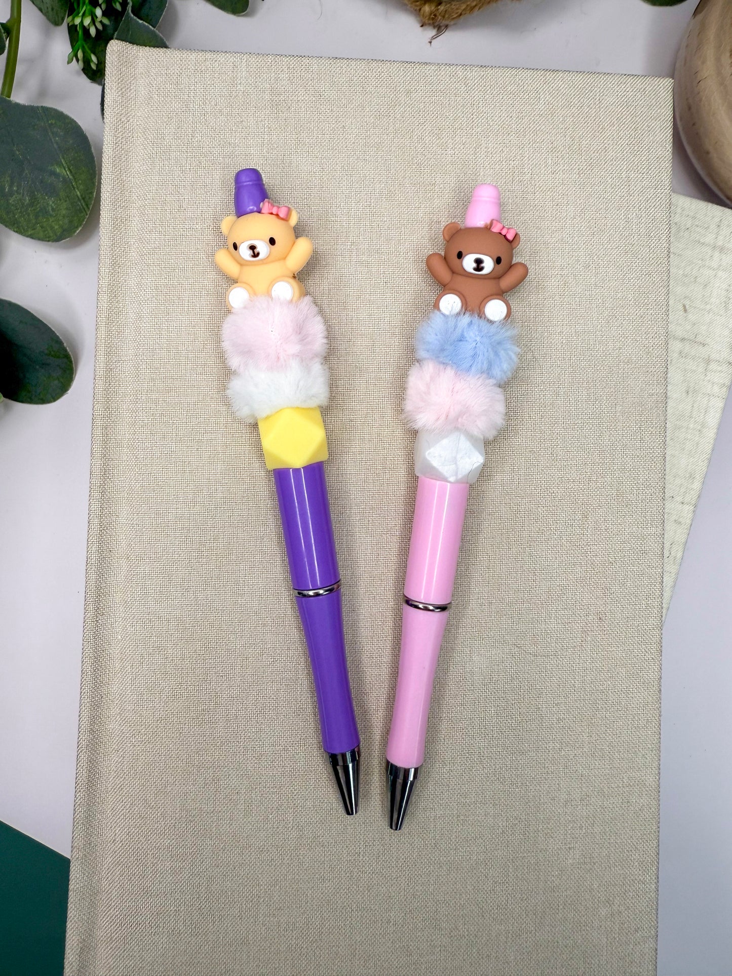 Two colorful pens with teddy bear decorations are placed on a beige surface.