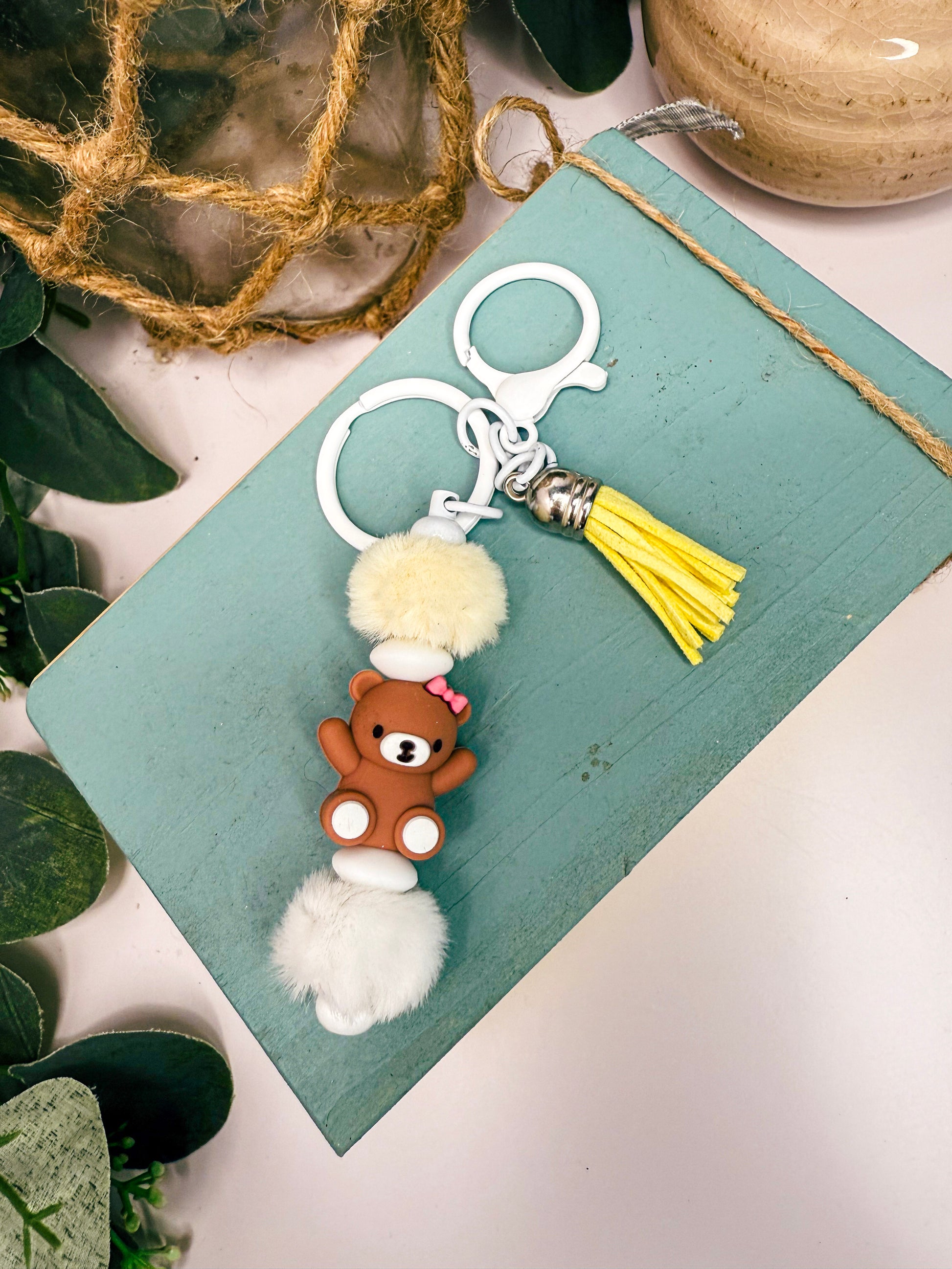 A keychain with a brown teddy bear charm and a yellow tassel is placed on a light blue surface, surrounded by greenery.