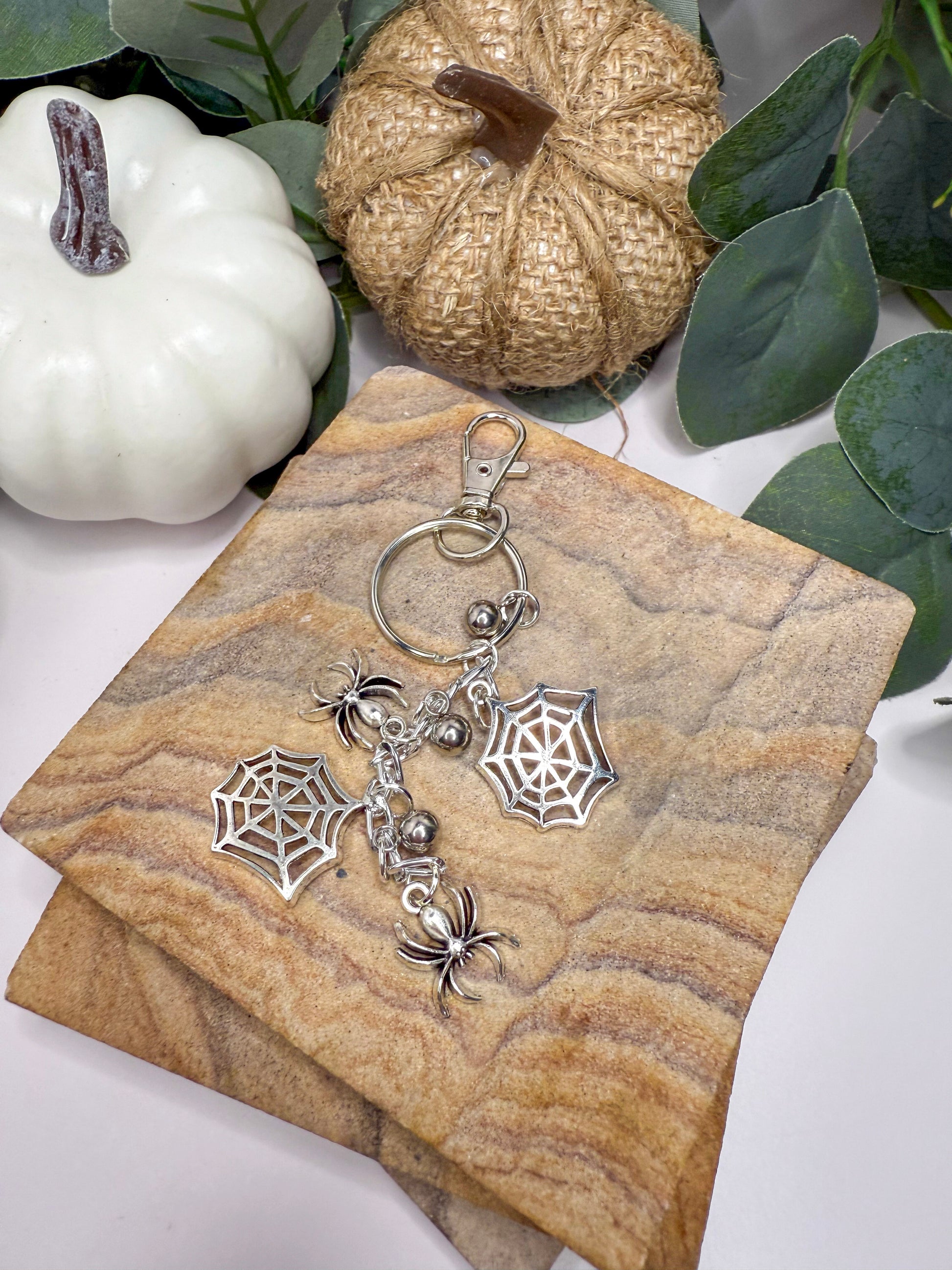 a keychain with a spider design, placed on a textured surface. There are also small white pumpkins and some greenery in the background.