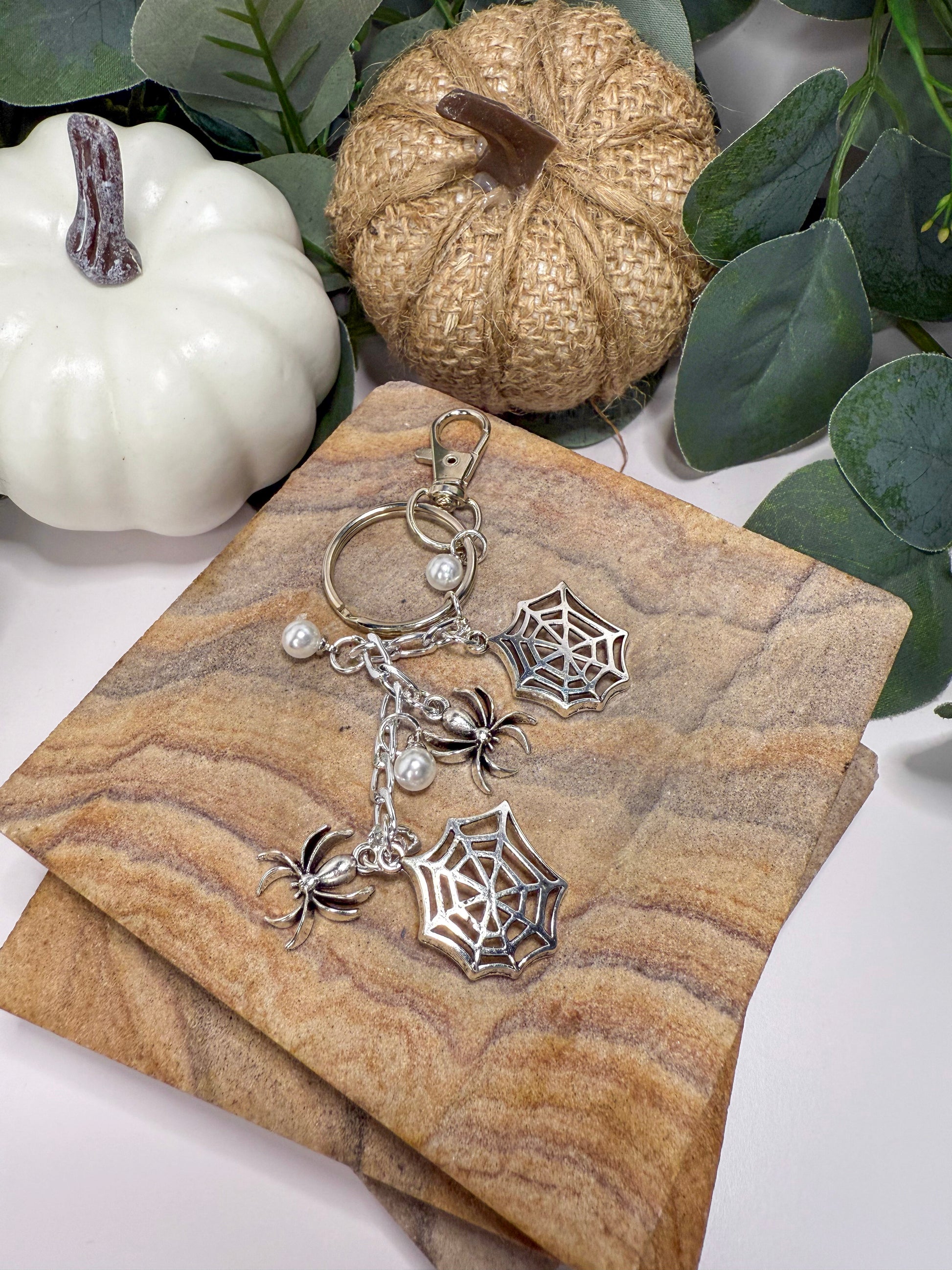 a keychain with a spider web design, placed on a wooden surface. The keychain has a chain and two small spider web charms. There are also two small white pumpkins and some greenery in the background.