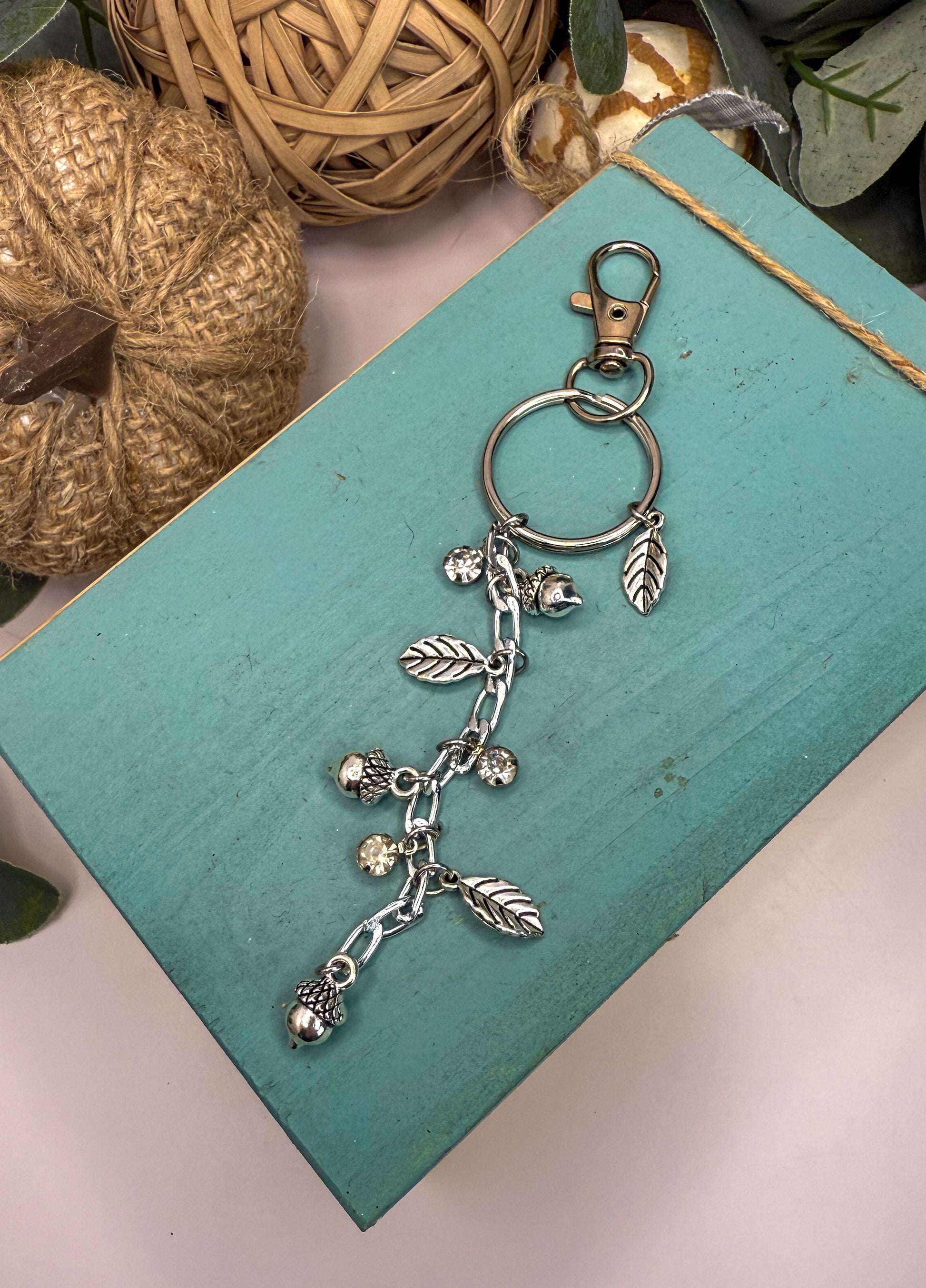 A silver keychain with a charm and leaf design rests on a blue surface, surrounded by other decorative items.