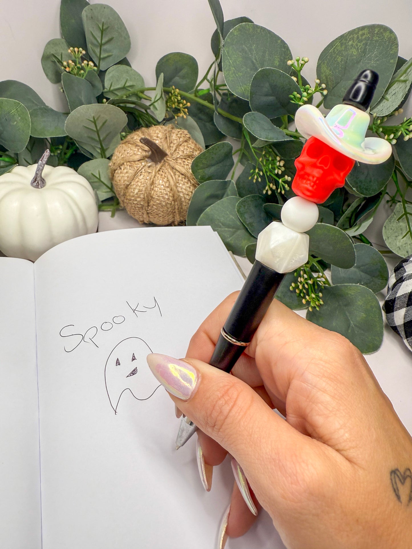 A hand holding a pen with a ghost drawing on a notebook, surrounded by Halloween decorations and greenery.